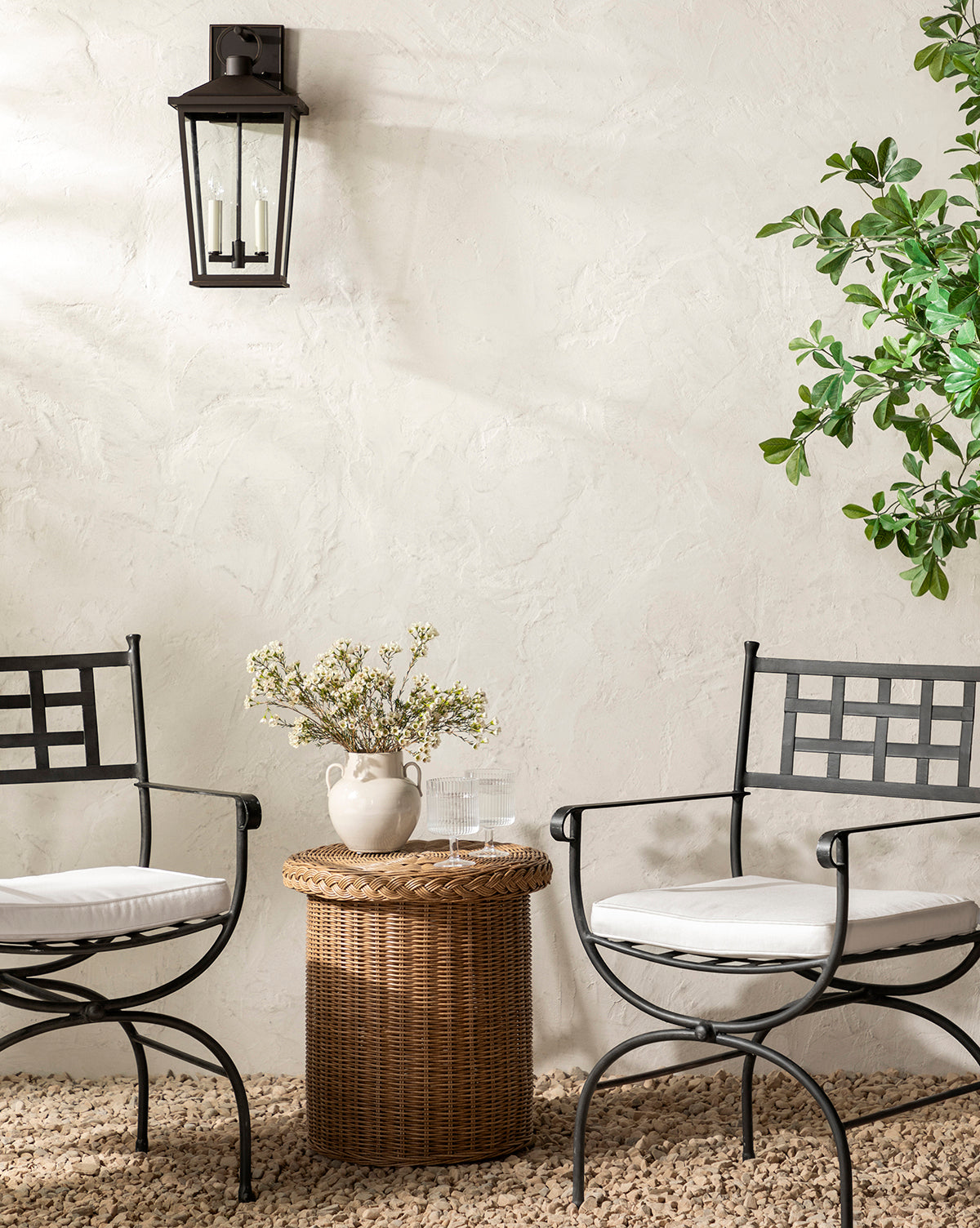 Two black metal chairs with white cushions invite outdoor lounging on a pebbled floor, facing the weather-resistant Haviland Side Table topped with flowers and water. A leafy plant and black wall lantern accent the neutral textured wall, by Vendixs