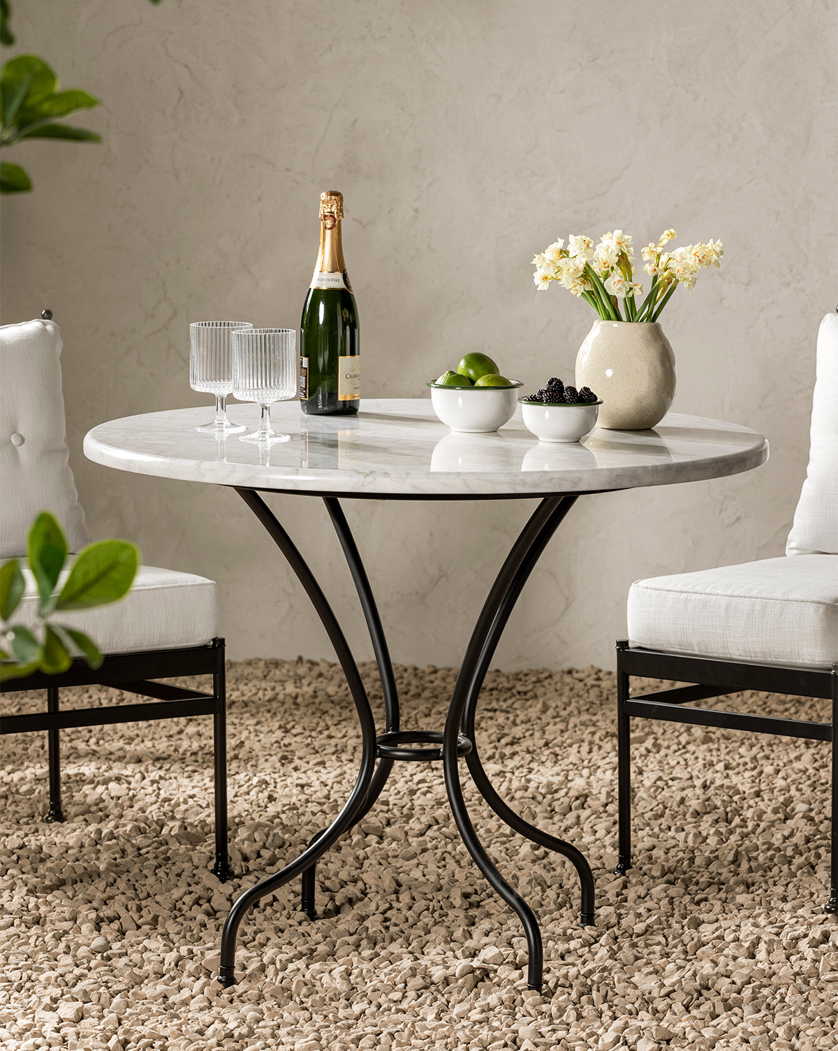 A round marble table with black curved legs stands on a gravel floor. On the table are two glasses, a bottle of champagne, a vase of daffodils, and Delia Bowls (Set of 3) filled with limes and blackberries. Two white cushioned chairs flank the table, by Vendixs