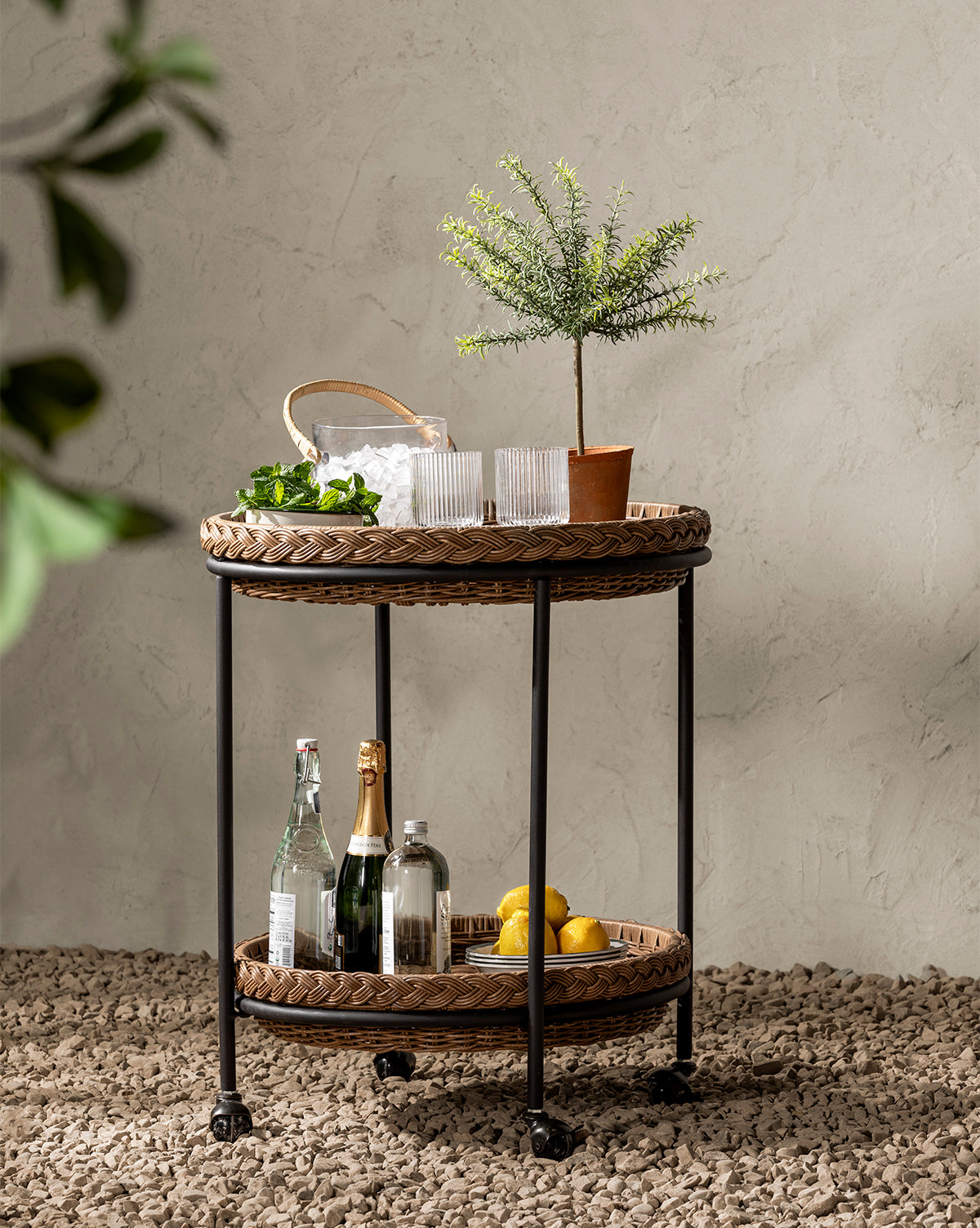 The Haviland Bar Cart, a weather-resistant wicker cart on wheels, sits on pebbles. Its top shelf holds a potted plant, glasses, and an ice basket; the lower shelf displays bottles and lemons—ideal for chic outdoor entertaining. A leafy plant is in front, by Vendixs