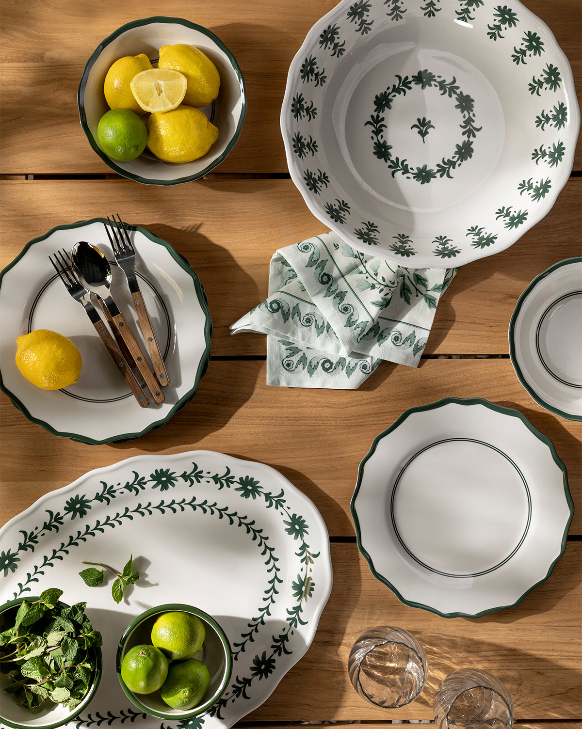 A wooden tabletop displays white dishes with a green floral motif, bowls of lemons and limes, herb sprigs, cutlery, napkin, glasses, and the Alba Melamine Serving Bowl with a scalloped edge, by Vendixs