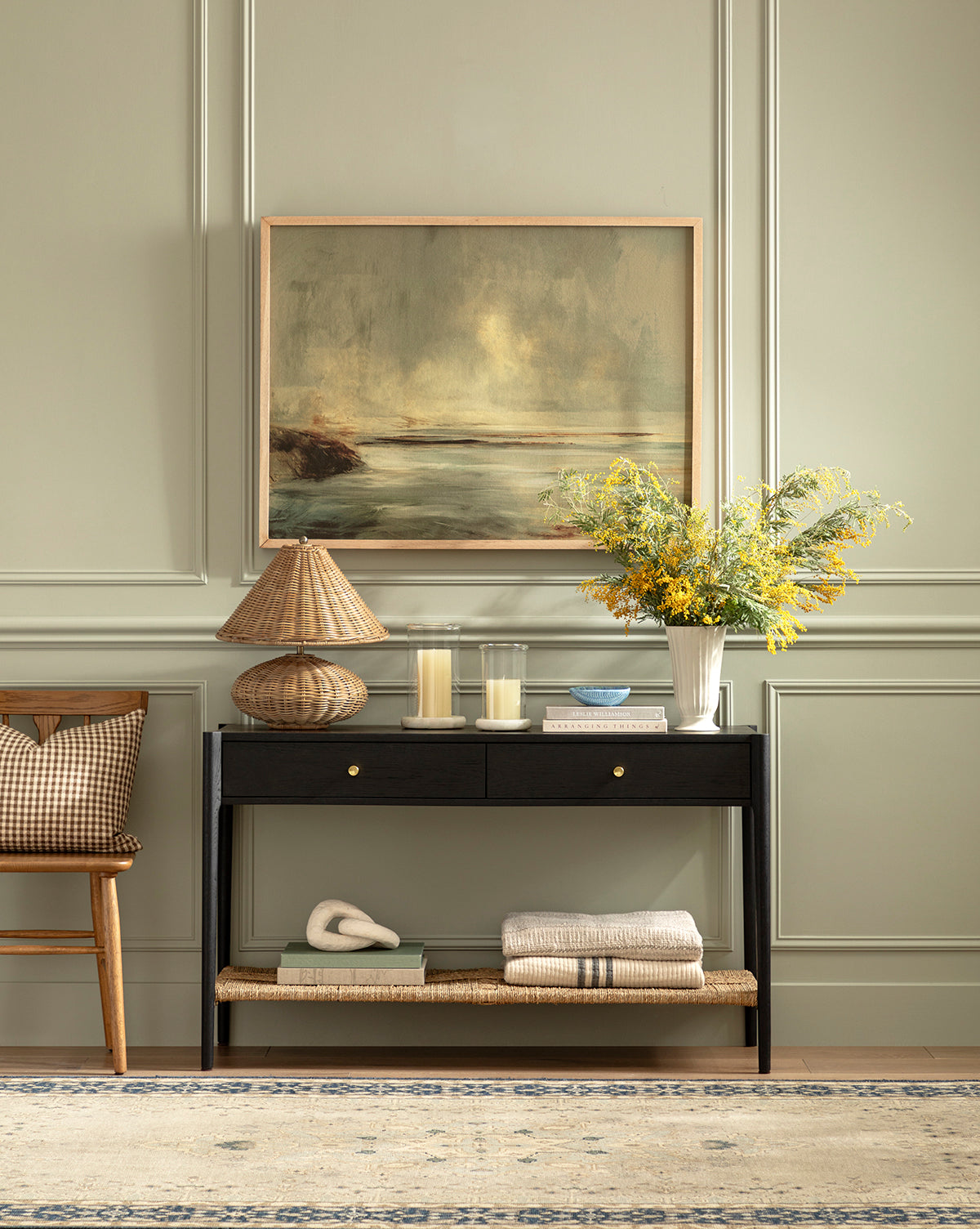 The Dana 2-Drawer Console, topped with a lamp, candles, and a yellow flower vase, stands against a pale green paneled wall beneath a framed landscape—creating an elegant entryway beside a chair and folded blankets by Vendixs