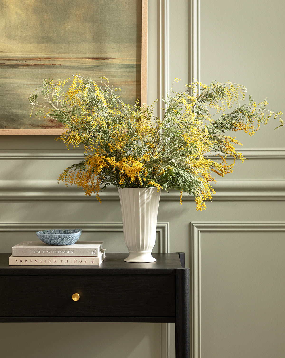 A Delphi Footed Vase filled with yellow and green wildflowers sits on a dark wood table beside a small bowl and stacked books, in front of a sage green paneled wall and framed painting, by Vendixs