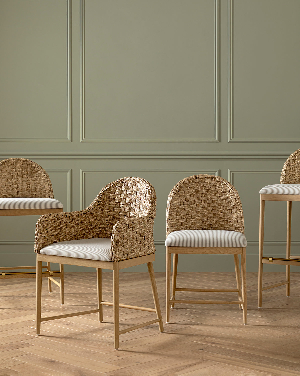 Four Molly Dining Chairs with woven rattan seats and solid oak legs are set on a wooden floor before a sage green paneled wall; two feature armrests and two are taller bar stools, by Vendixs