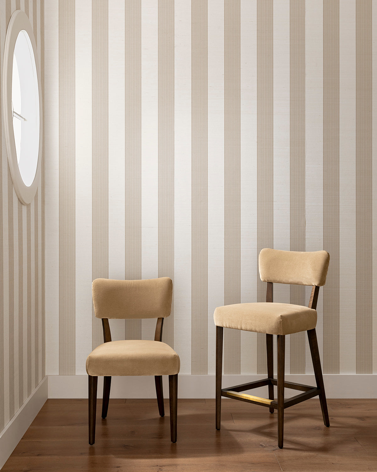 Two beige upholstered chairs with dark wooden legs sit on a wooden floor in the corner, next to the Makerspalm Etienne Stool (Ready to Ship). Behind them is a vertically striped beige and white wall with a round window on the left. Vendixs