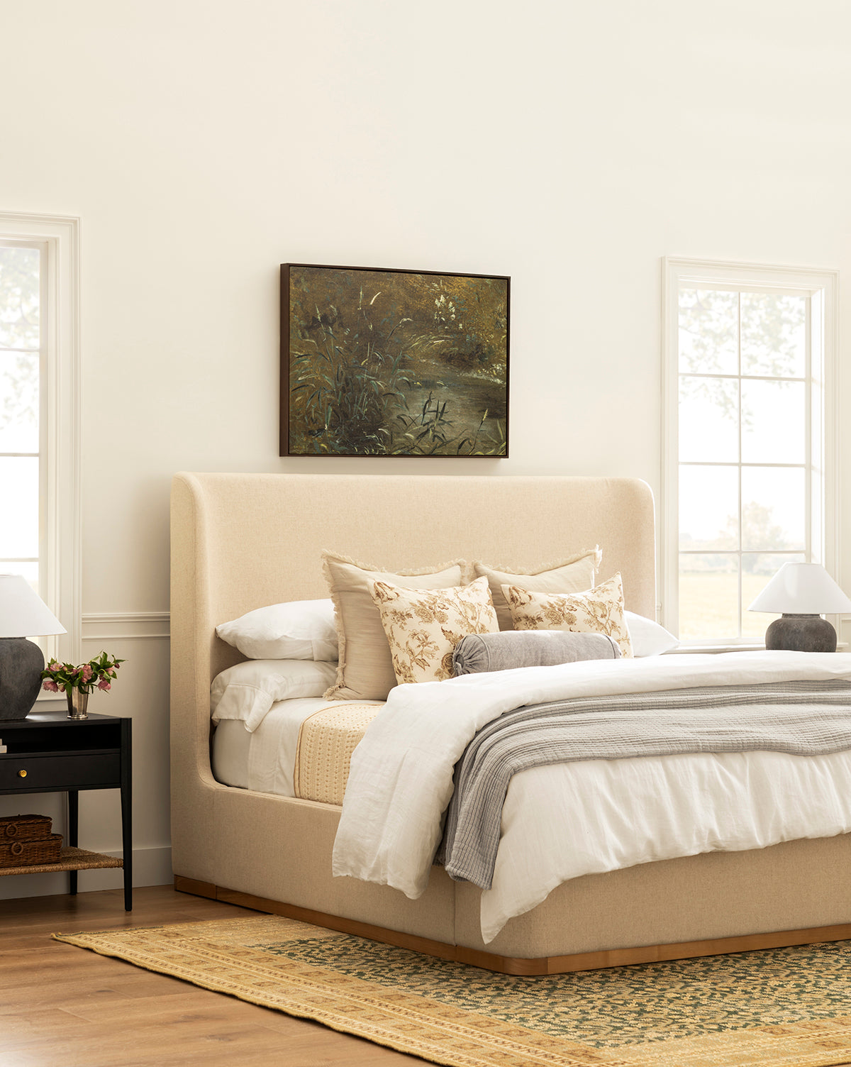 A cozy bedroom with the Denning Upholstered Bed by Makerspalm in beige, layered neutral bedding, floral pillows, two black nightstands with lamps, a yellow woven rug, large windows, and a nature painting above the bed. Vendixs