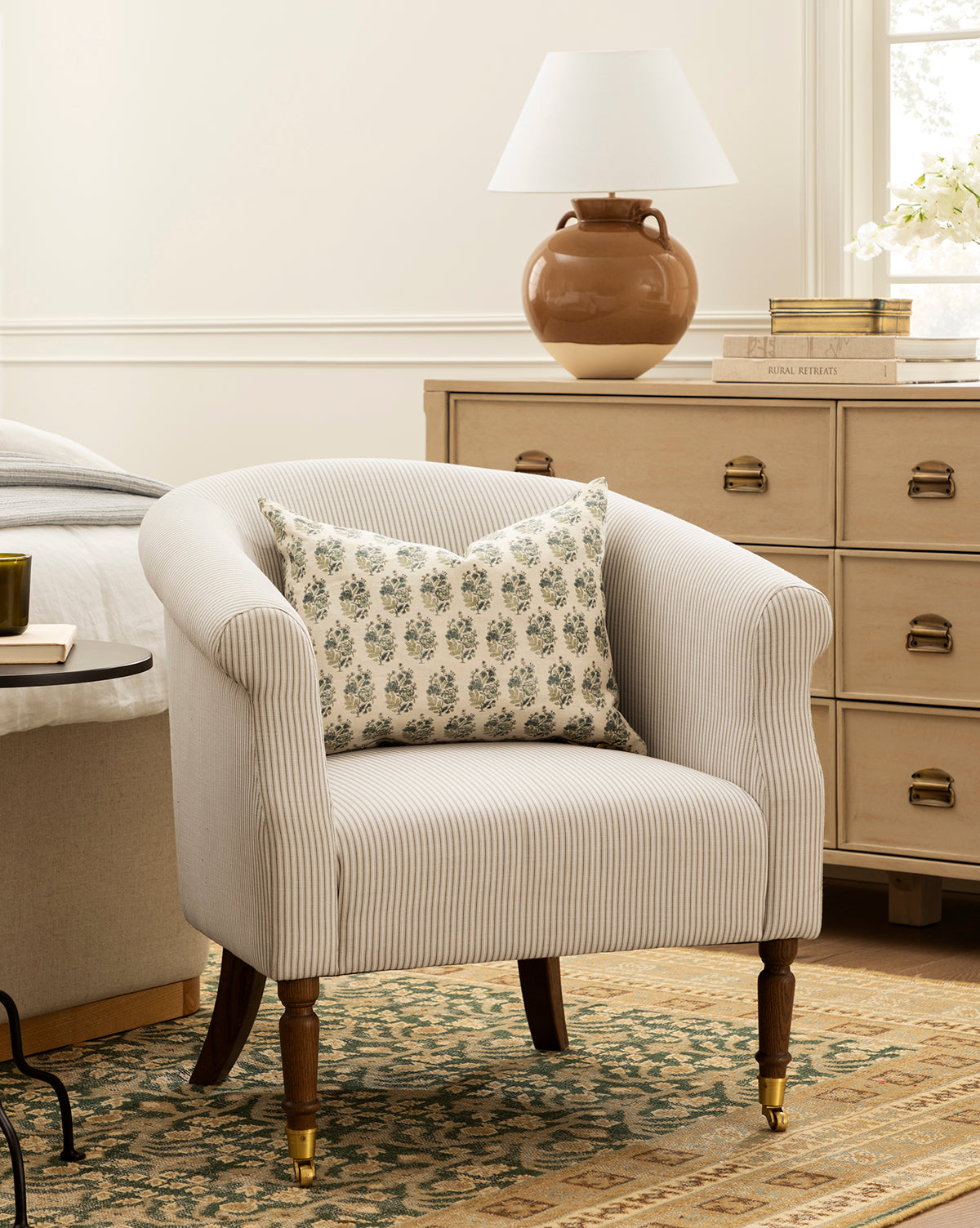 The Makerspalm Clemence Lounge Chair with a patterned pillow sits on a patterned rug beside a wooden dresser topped with a ceramic lamp, books, and flowers in a cozy, well-lit bedroom, Vendixs