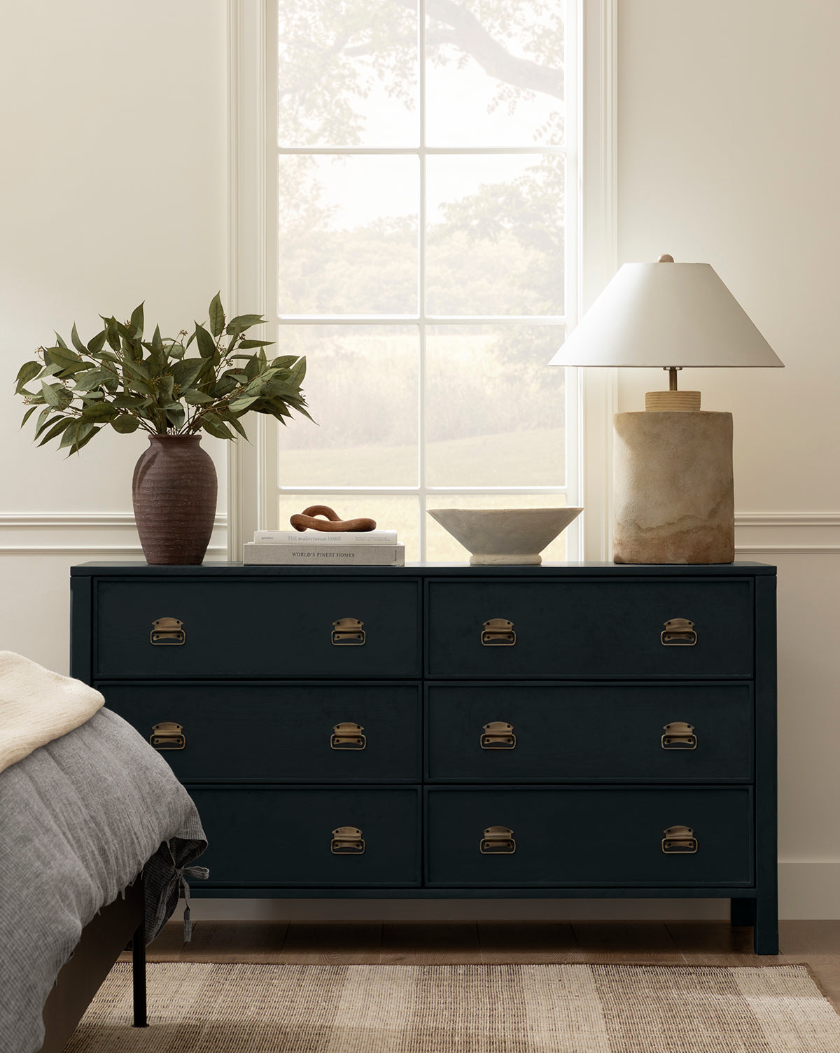 A navy blue dresser with brass handles sits beneath a large window. On top are a brown vase with green leaves, a white bowl, a small tray, and the Samala Lamp with a white shade. Part of a bed is visible at left by Vendixs