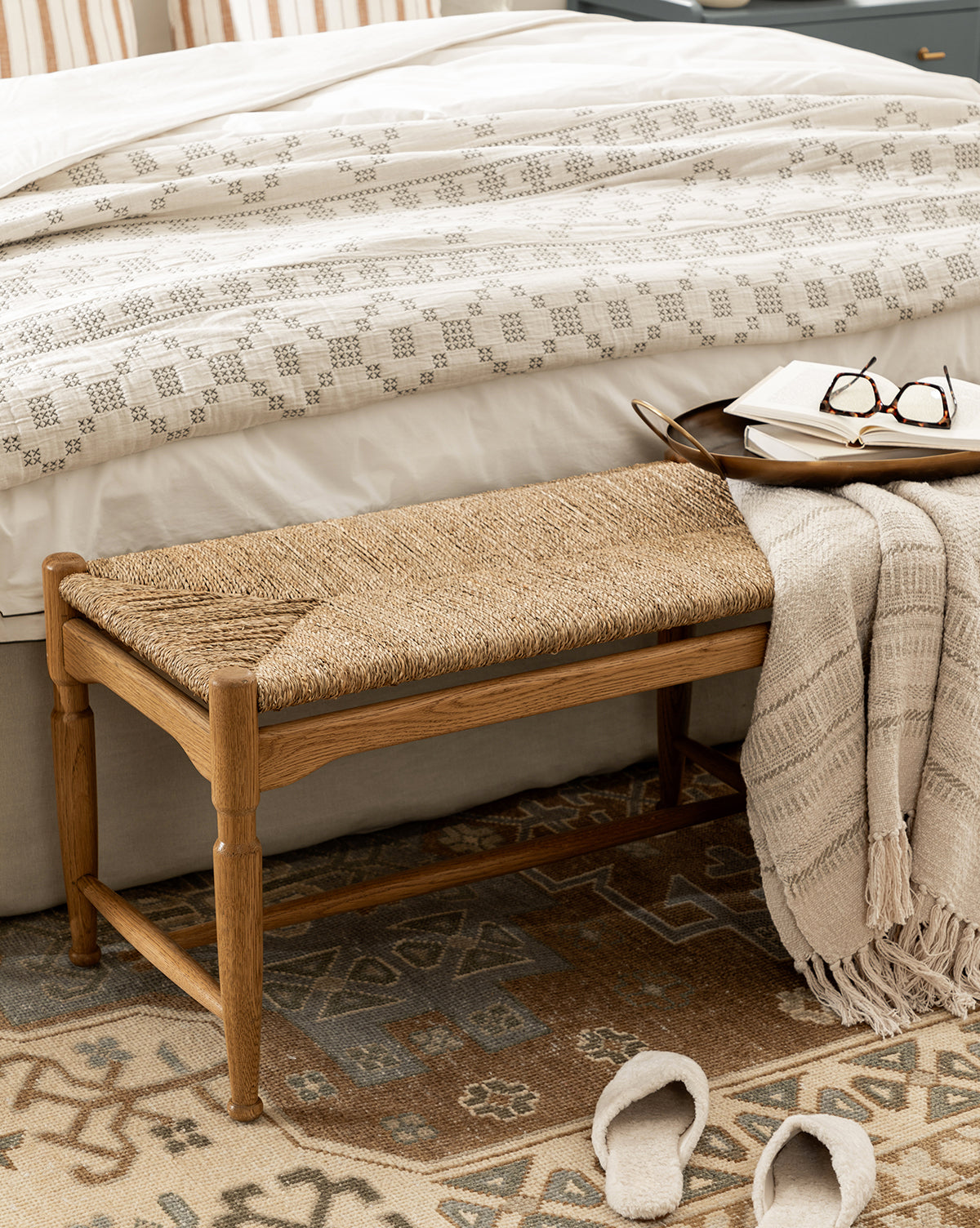 The Phipps Woven Bench with a seagrass seat and beige throw blanket sits at the foot of a bed with patterned bedding. A tray with a book and glasses rests on the bench, while slippers sit nearby on a patterned rug, by Vendixs