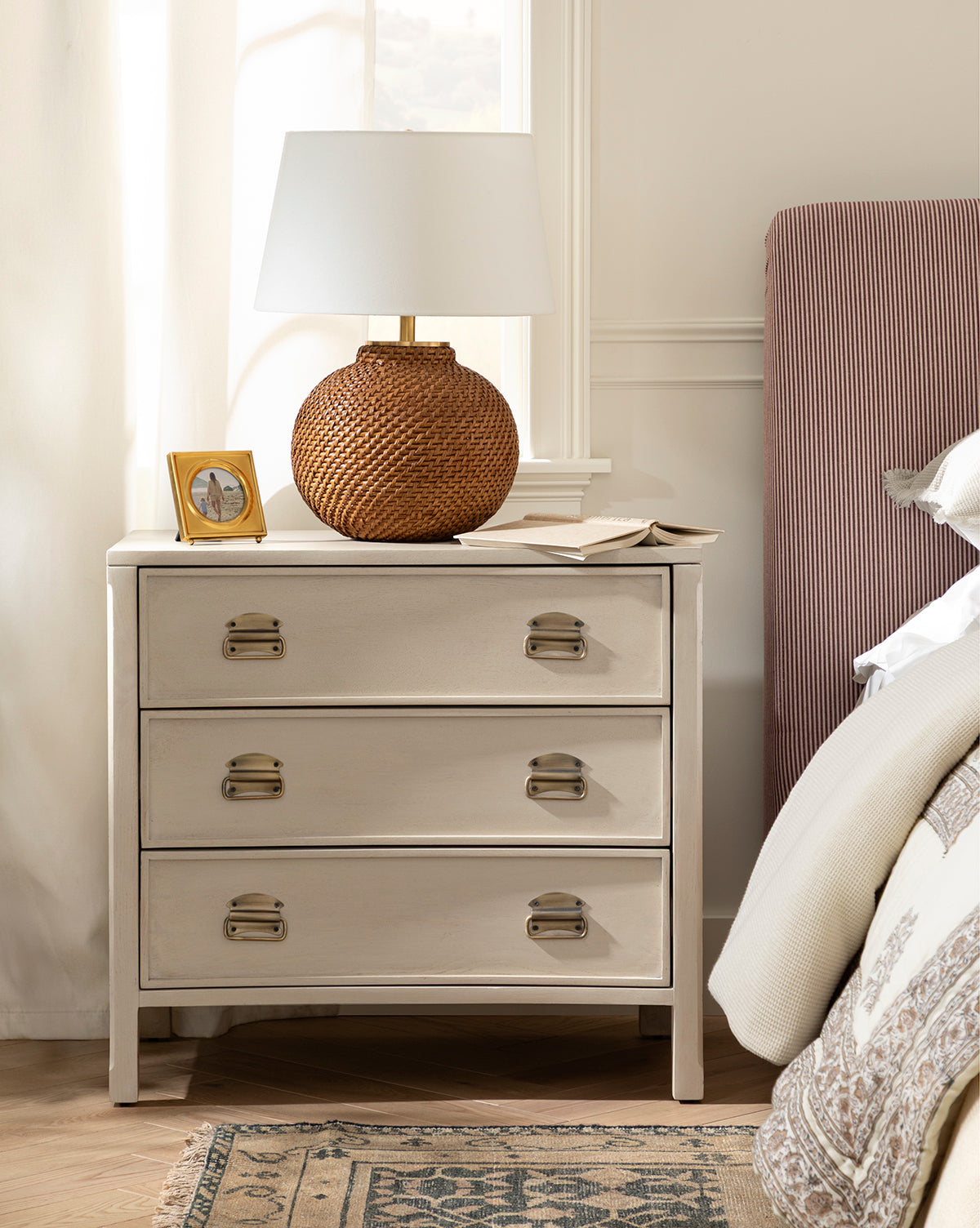 A beige nightstand with three drawers holds the Avedon Table Lamp with a natural rattan base and white linen shade, a small framed photo, and an open book. Beside it sits a bed dressed in neutral bedding and topped with a red-striped upholstered headboard by Vendixs
