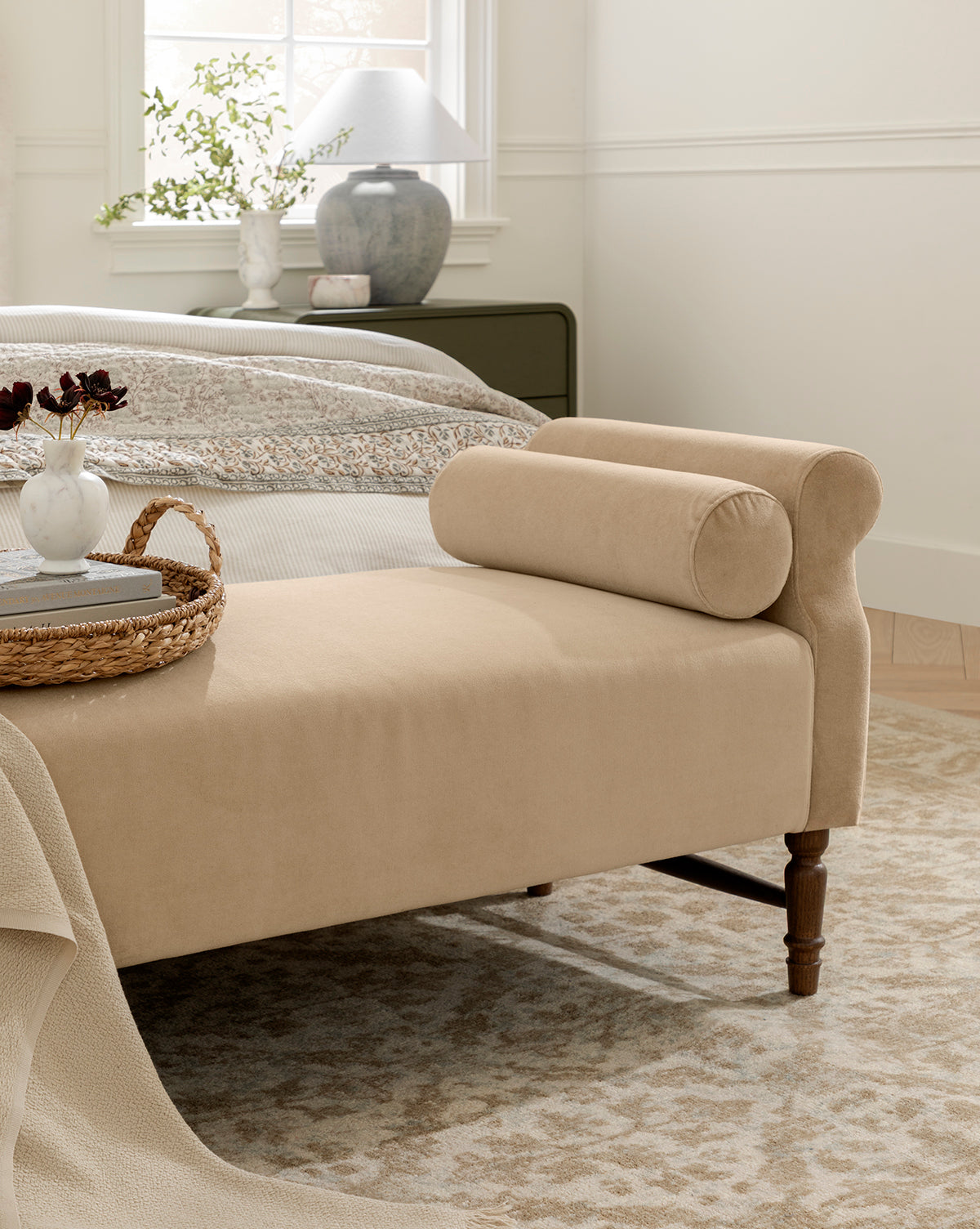 A beige upholstered Asher Bench sits at the foot of a neatly made bed in a cozy bedroom, featuring a cylindrical bolster pillow and topped with a woven tray, vase, and decor. Sunlight pours in from the window behind the bed, by Vendixs
