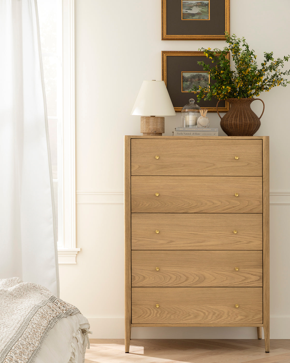 A light wood dresser with four drawers and gold knobs stands by a white wall. On top are a brown vase with green branches, the Bronte Table Lamp, and framed art above. Sunlight streams in from a nearby window by Vendixs