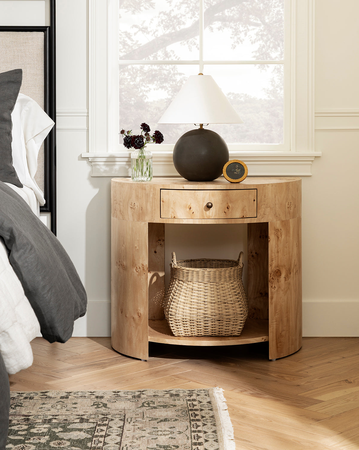 A round wooden nightstand with a drawer and open shelf holds the Boyce Woven Basket below. On top, there’s a lamp, small clock, and a vase with dark flowers, all set by a window next to a bed with gray and white bedding by McGee & Co.