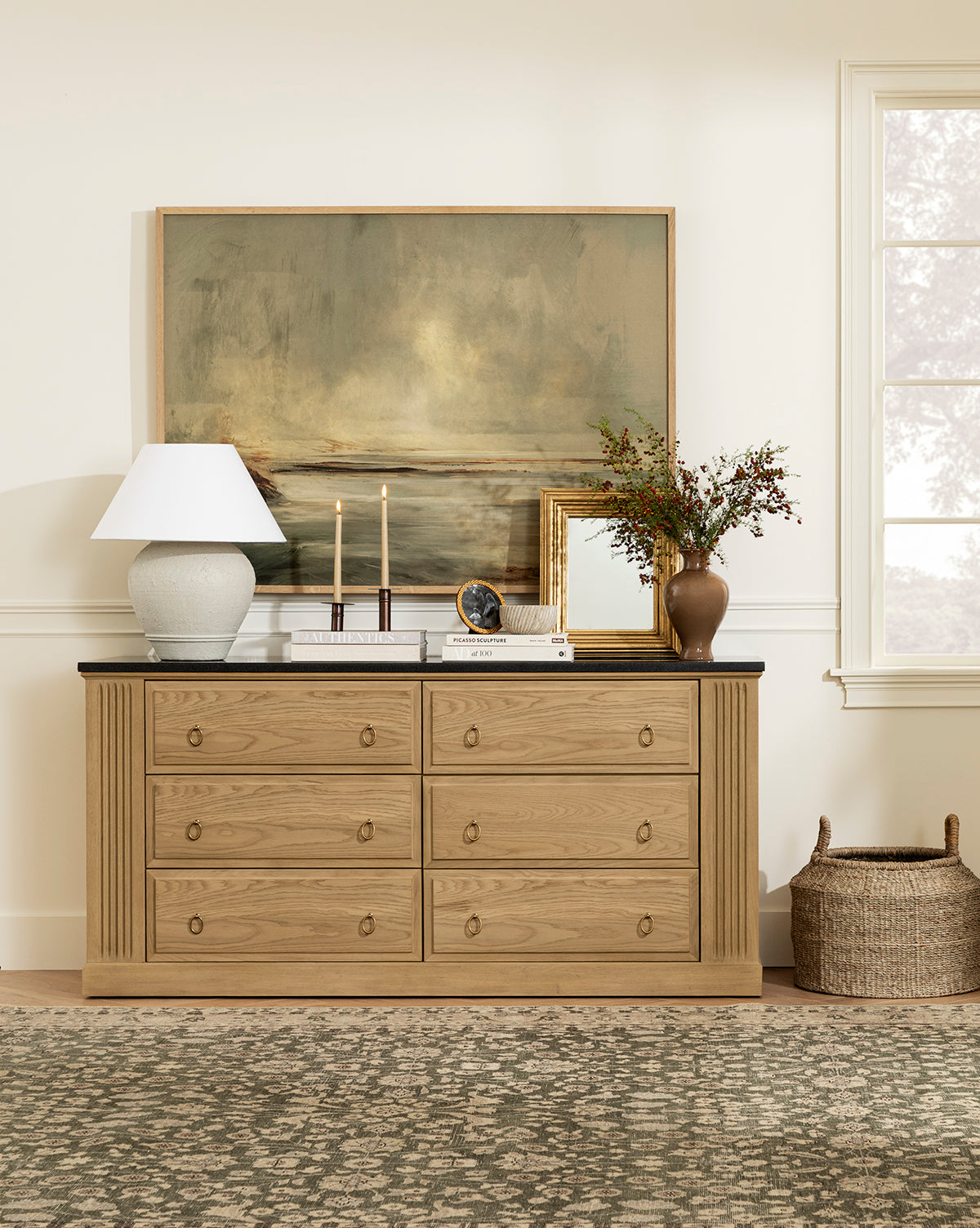 A wooden dresser with six drawers holds the Abbott Ceramic Table Lamp with a white linen shade, two lit candles, a clock, framed art, and a brown vase with greenery. Above leans a large landscape painting; a basket sits on the floor nearby, by Vendixs