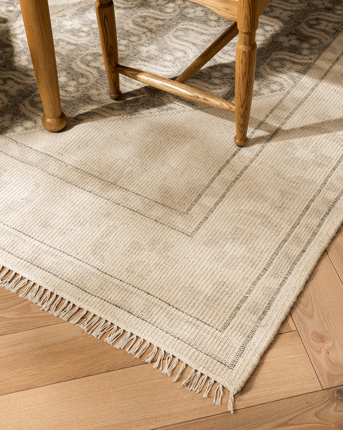 A close-up of the Elin Hand-Knotted Wool Rug in light beige, featuring artisan details, a subtle striped border, and fringed edges on a wooden floor, partially beneath a wooden chair, by Vendixs