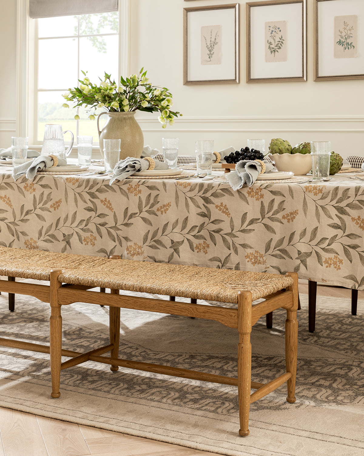 A dining table with a floral tablecloth is set with plates, glasses, napkins, and a ceramic jug holding green branches. The room features the Phipps Woven Bench and framed botanical prints for an elegant, neutral-toned look by Vendixs