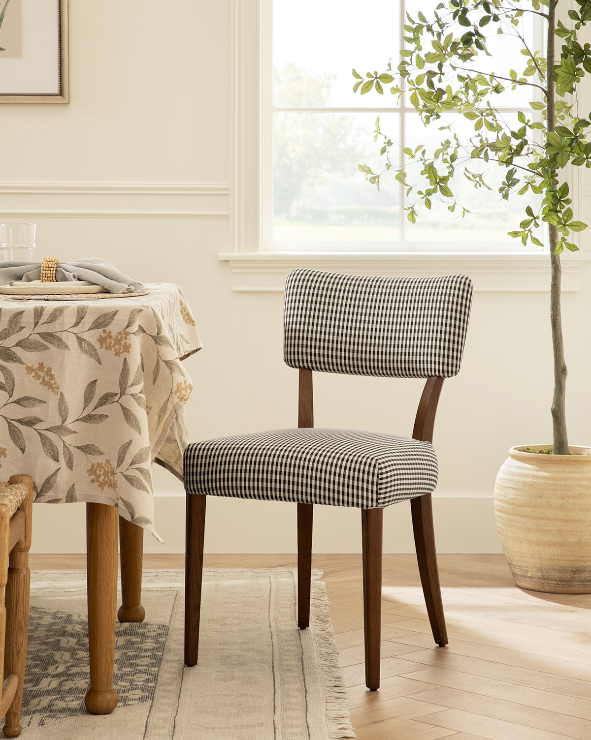 A dining room with a wooden table draped in a floral tablecloth, an Etienne Dining Chair with black and white checkered upholstery, a large potted plant, and sunlight pouring through the window behind by McGee & Co.