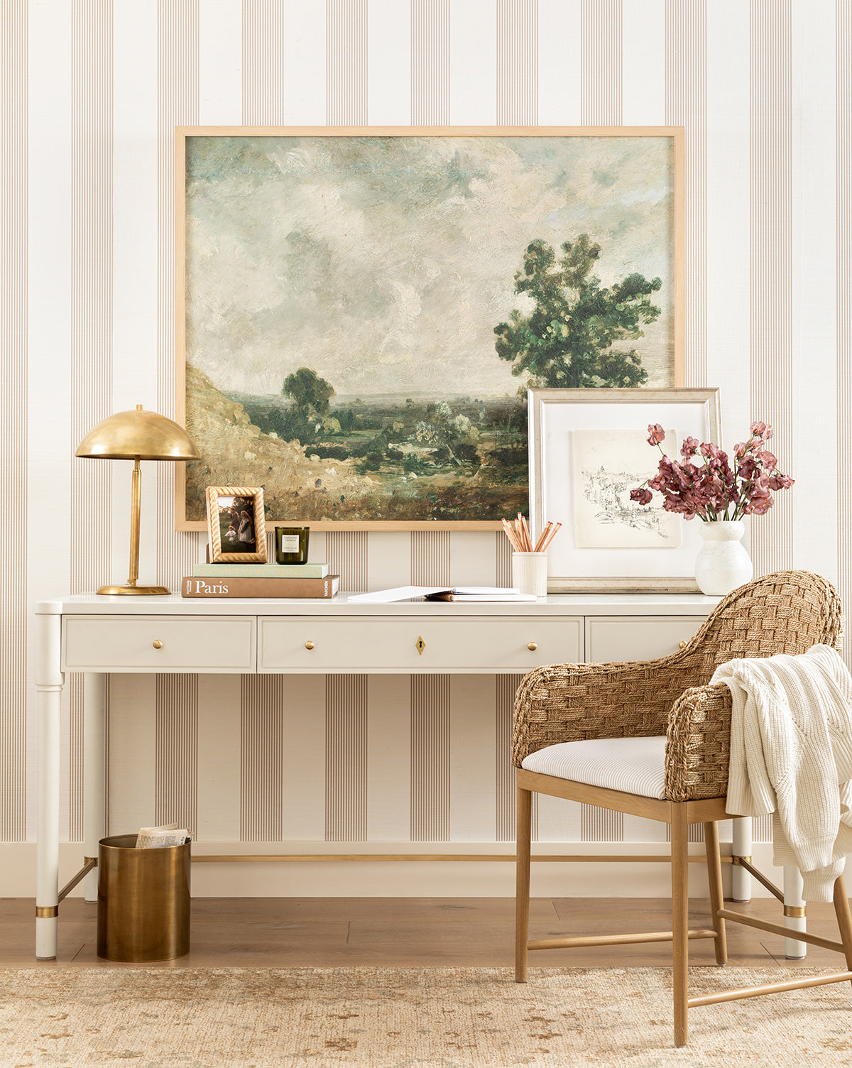 A stylish workspace with a white desk, gold lamp, books, pink flowers in a vase, and a wicker chair with cushion. The striped wallpaper highlights the English Meadows giclee on canvas wall art in a natural oak frame, by Vendixs