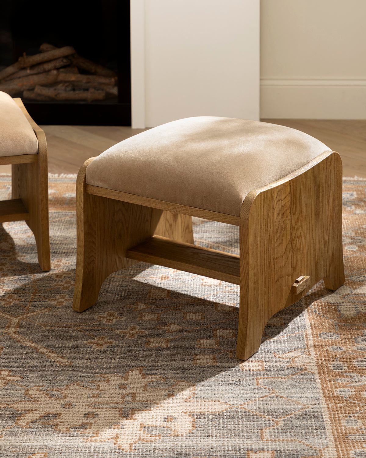 A Makerspalm Gable Ottoman in Drift Oak with a Fawn Velvet cushioned top rests on a patterned rug in a bright room featuring light walls and wood floors, Vendixs