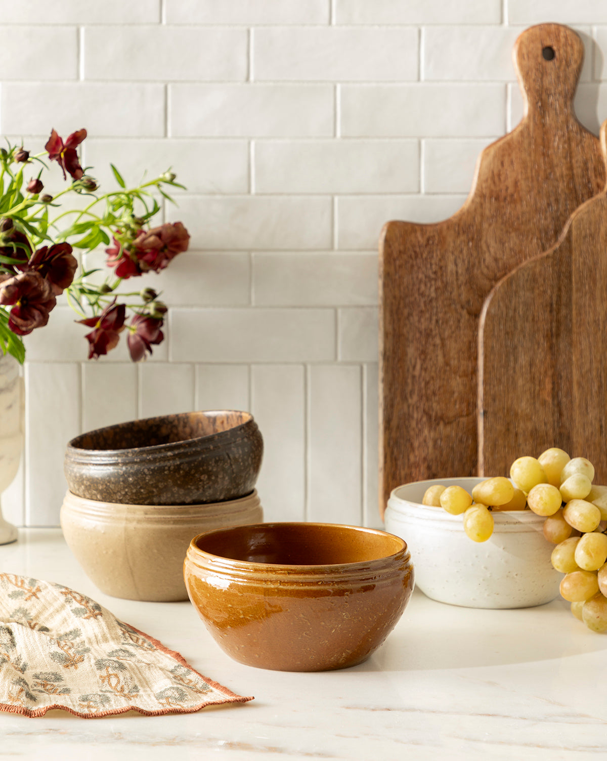 A set of four Mariella Stoneware Bowls in brown, beige, and white are displayed on a kitchen counter near grapes, wooden cutting boards, a patterned cloth, and a vase with dark red flowers against a white tile backsplash, by Vendixs