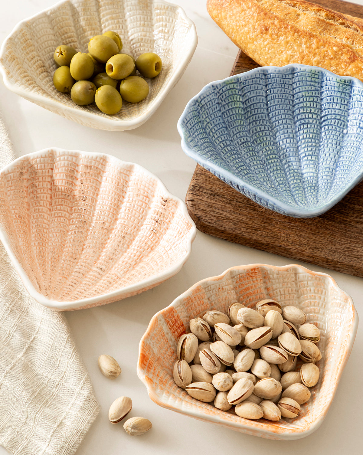 Four Aurelie Shell Shaped Bowls (Set of 4) in pastel tones hold green olives and pistachios. A baguette sits on a wooden board nearby, with a cloth napkin partially visible on the left, by Vendixs