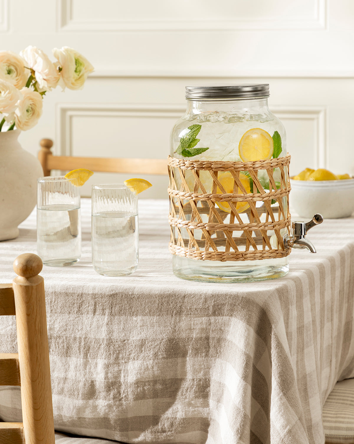 The Glass Beverage Dispenser with Woven Sleeve, filled with lemon slices and mint, sits on a striped tablecloth beside two glasses of water, a bowl of lemons, and a vase of flowers in the background, by Vendixs