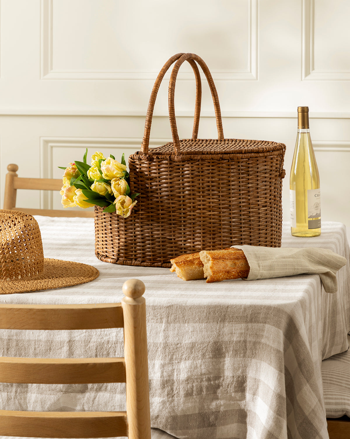 The Rattan Wicker Picnic Basket sits on a striped tablecloth next to a straw hat, baguette, and white wine. Surrounded by wooden chairs in a softly lit room, it offers an inviting and stylish storage solution for gatherings by Vendixs