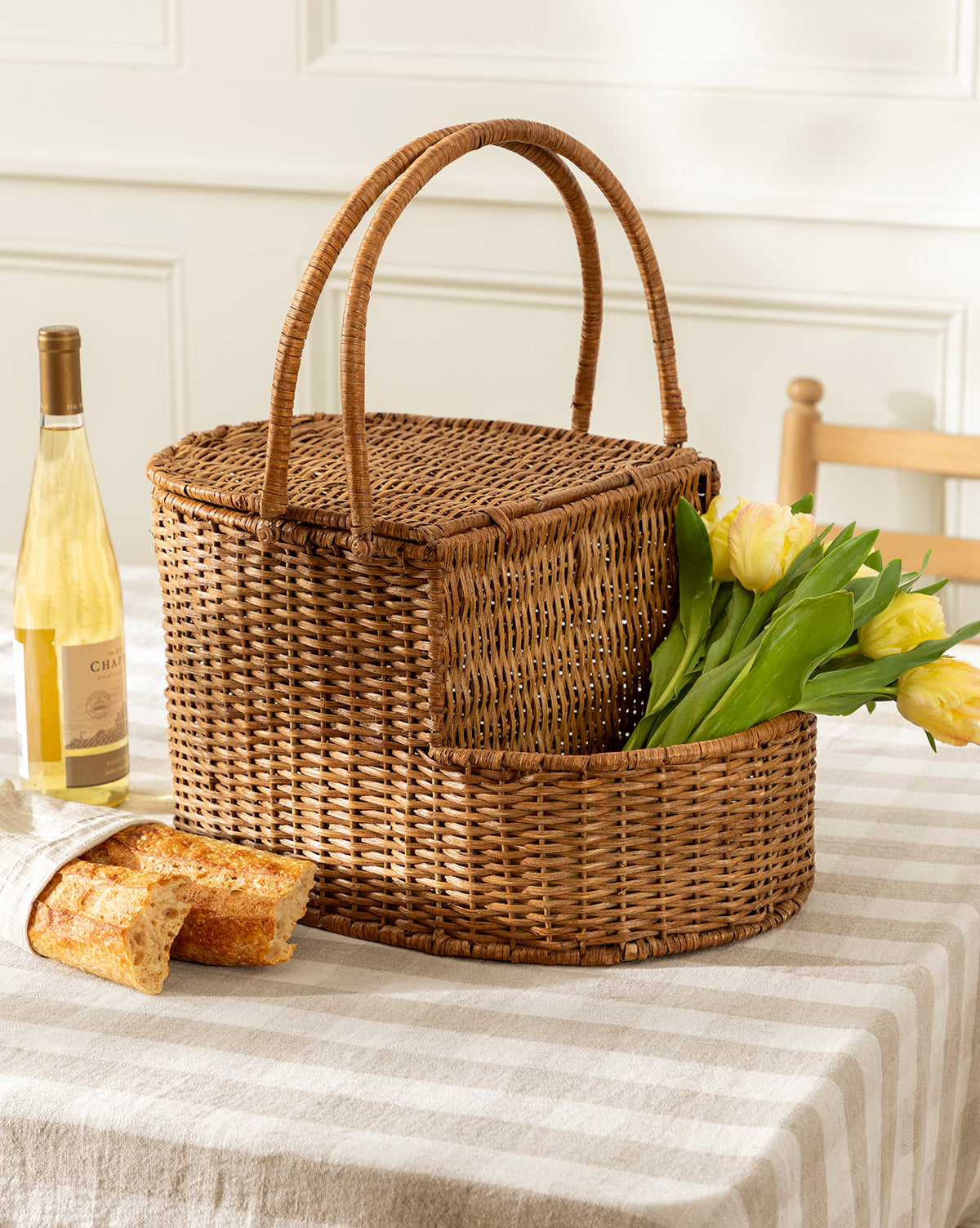 A Rattan Wicker Picnic Basket rests on a striped tablecloth beside a bottle of white wine and a baguette, with a wooden chair and white paneled wall behind, creating an inviting setting for this charming storage piece, by Vendixs