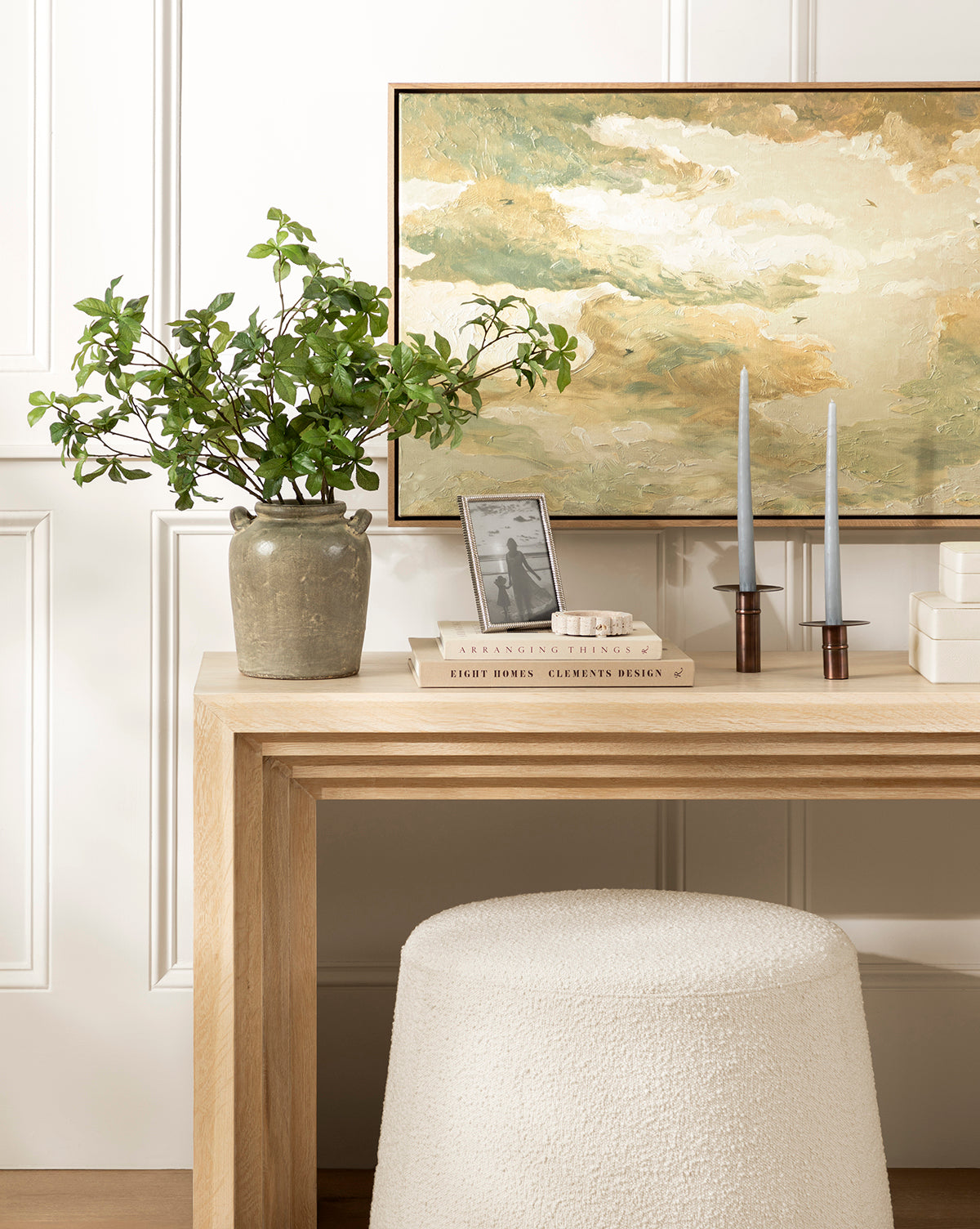 A light wood console table displays a potted leafy plant, Swallows at Dusk wall art in a white oak frame, two blue taper candles in holders, stacked books, and a small photo frame; a textured white stool sits beneath, by Vendixs