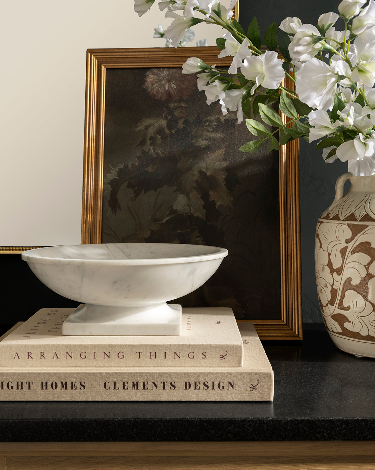 The Tayva Bowl sits atop two stacked design books on a dark countertop, with a framed floral painting and a ceramic vase of white flowers behind it to complete the charming display, by Vendixs