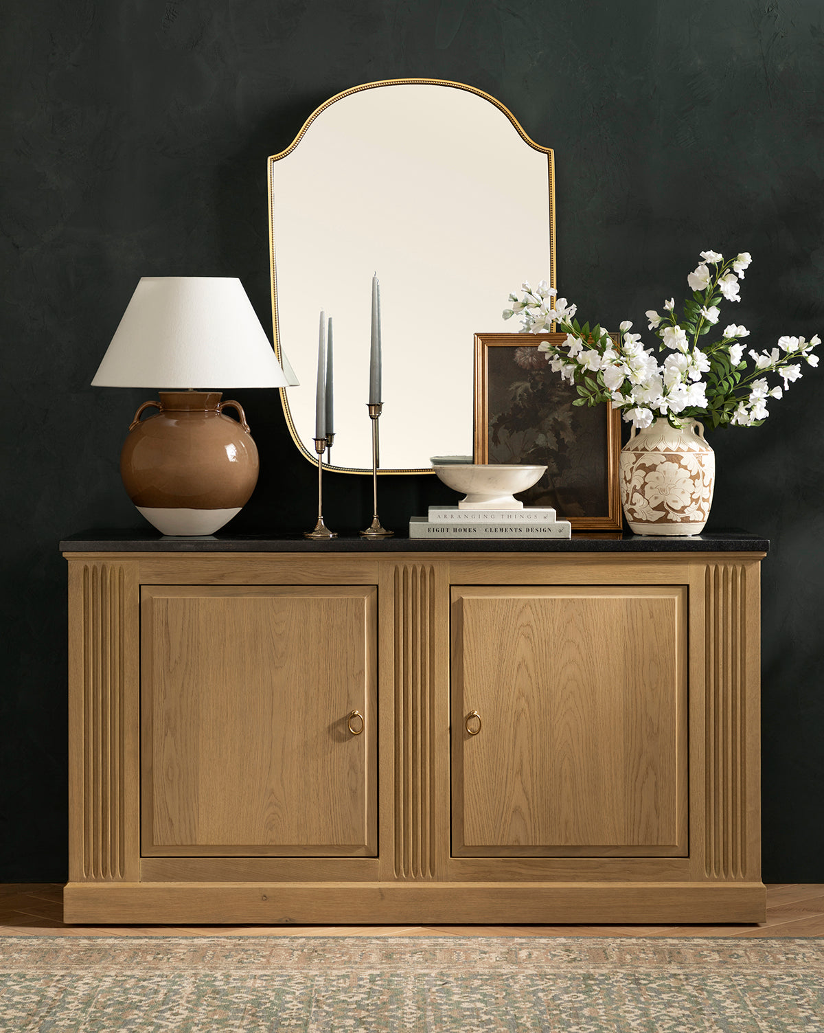 The Ambrose Sideboard by YANG CHENG WOODEN INDUSTRIES INTERNATIONAL (VIETNAM) CO LTD features two doors and is styled with a brown lamp, candles, books, a bowl, framed art, a white flower vase, and a gold-edged mirror above. Vendixs