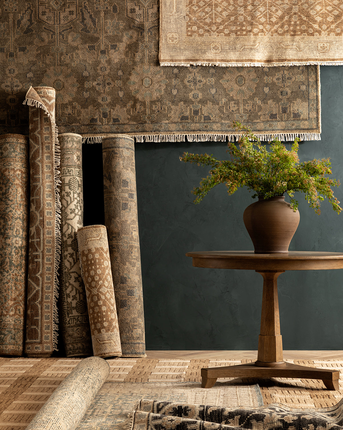 The Thames Hand-Knotted Rug in earthy tones is displayed against a dark wall, while a round wooden table with a brown vase of green foliage sits in front, creating an atmosphere of refined luxury home decor, by Vendixs