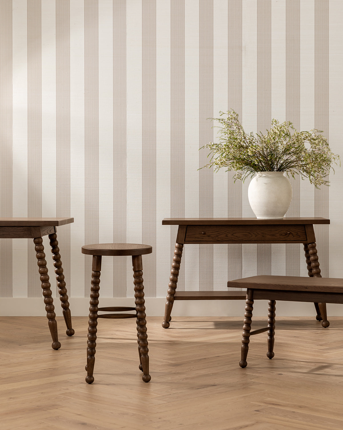 A minimalist room with light striped wallpaper, wooden furniture including the Heathcliff Bench with spindle legs, and a white vase with green foliage on a wooden table. The warm herringbone floor completes the look, by Vendixs