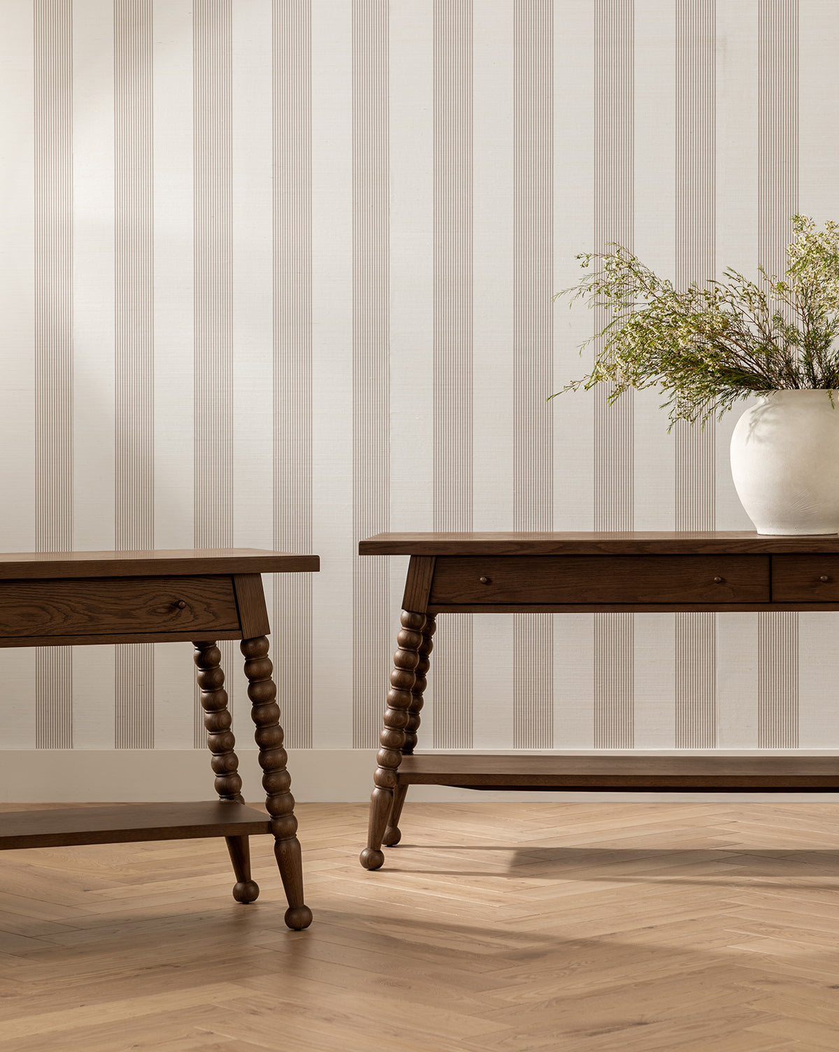 Two Heathcliff Console tables with spindle legs sit on a light herringbone wood floor near a beige and white striped wall. A large white vase with green foliage is placed on the right console, by Vendixs