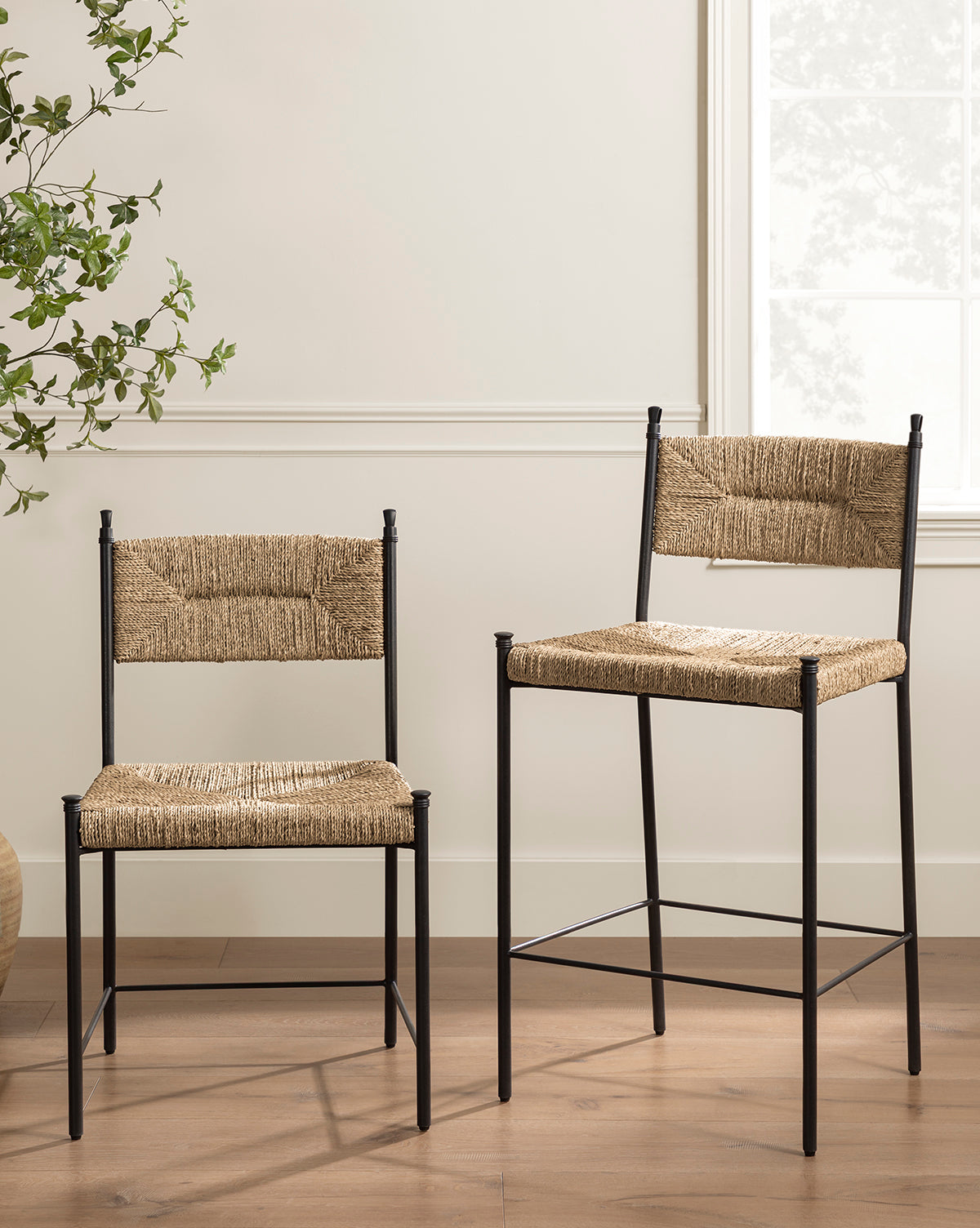 Two Makerspalm Peregrine Stools, featuring black metal frames and woven seagrass seats and backs, sit on a wooden floor near a window and leafy plant in a bright, modern room. One stool is taller for kitchen or dining use. Vendixs