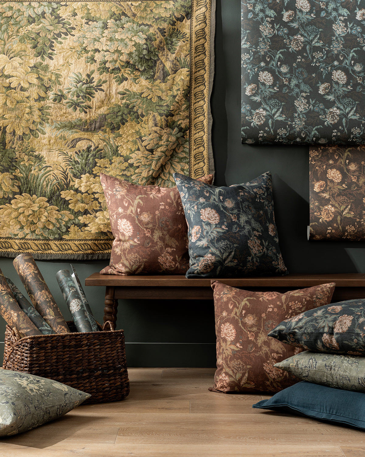 A cozy interior with painterly floral tapestries, blue and brown patterned cushions on a wooden bench, Hestercombe Floral Wallpaper by Makerspalm in a wicker basket, all set against a dark wall. Vendixs