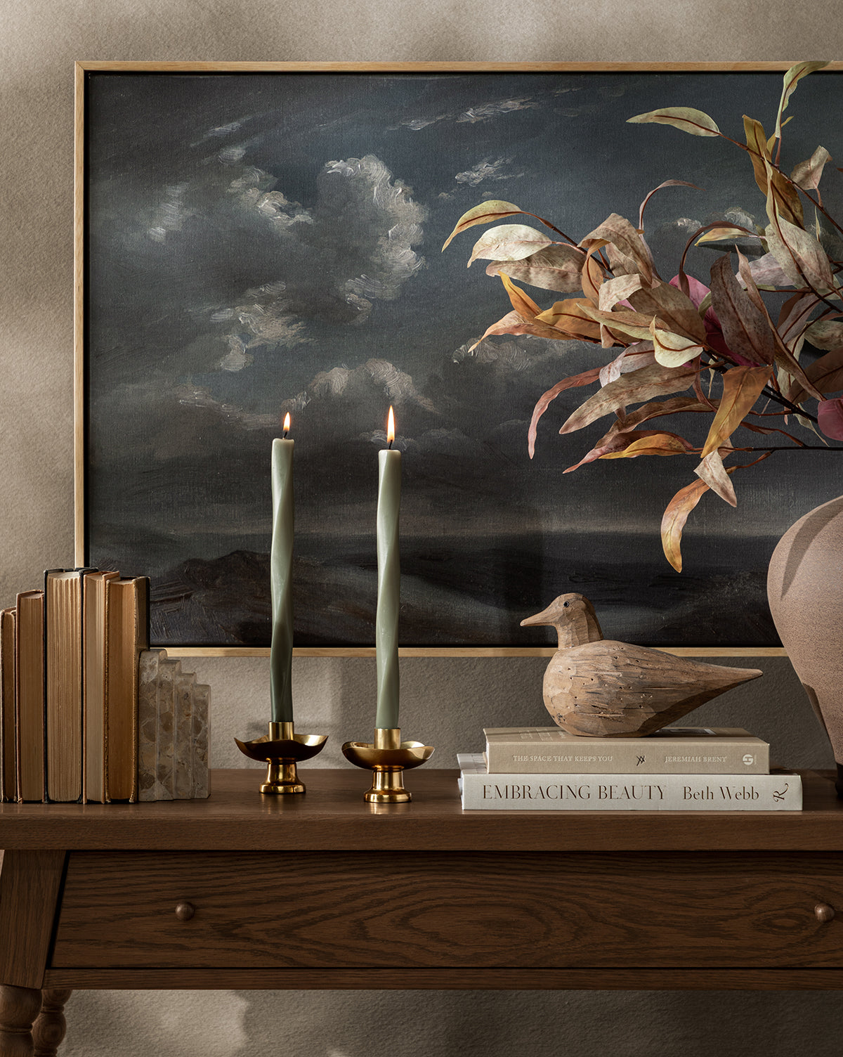 A wooden table holds two lit brass candle holders, a stack of books, a wooden bird figurine, and a large vase filled with Faux Purple Eucalyptus Stem, set against a backdrop of a cloudy sky painting, by Vendixs