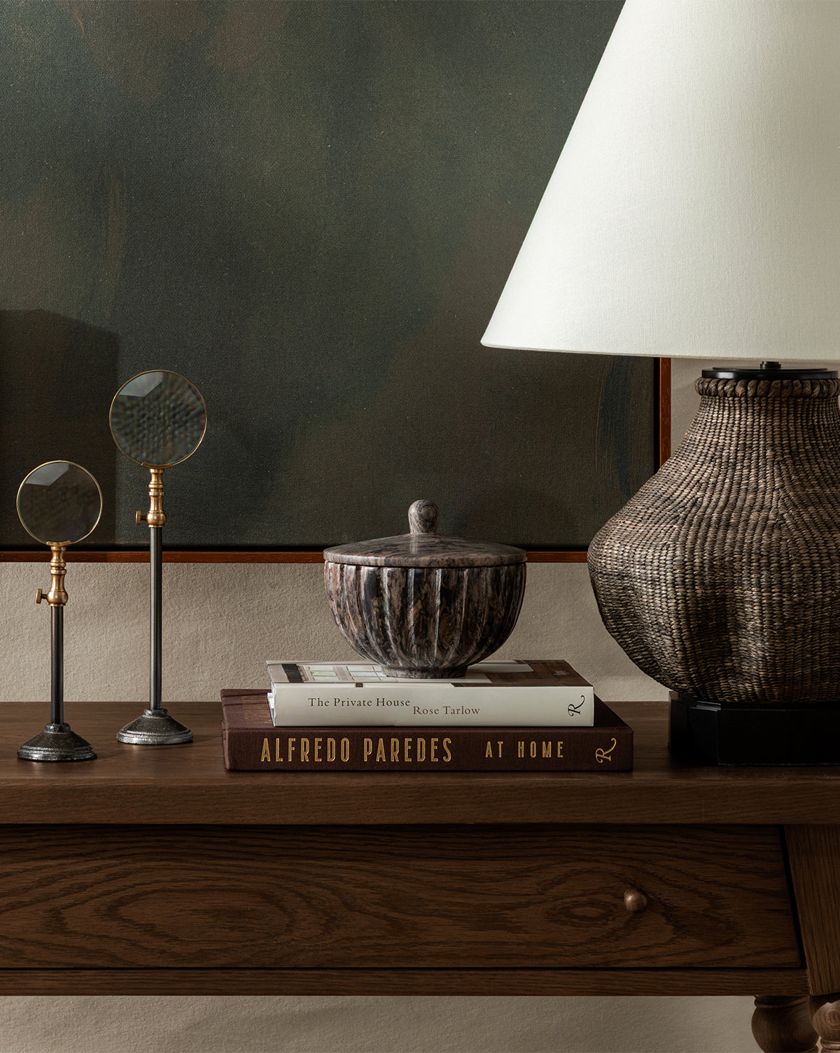 A wooden console table displays two stacked books, magnifying glasses on stands, a lidded bowl, and the Tahiti Table Lamp with a linen shade, all set before greenish wall art by Vendixs