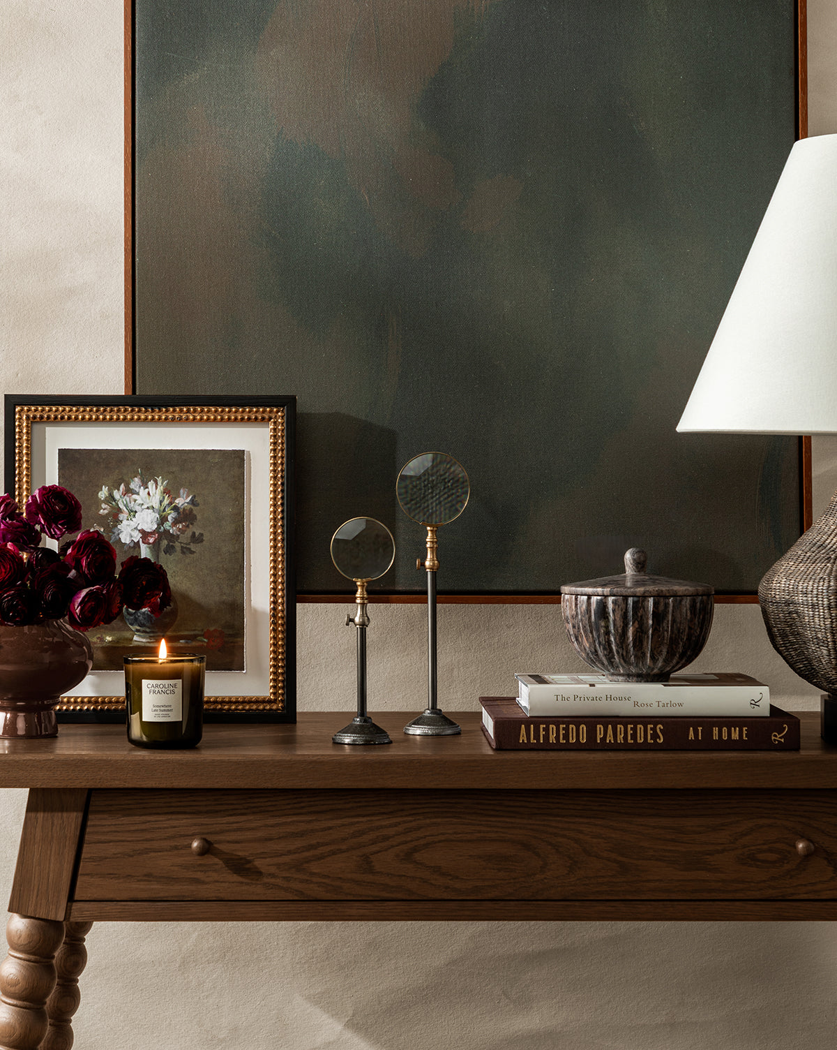 A wooden table with decor, including books, a lidded bowl, magnifying glasses, a candle, dark red flowers in a vase, and a framed floral painting sits beneath the striking abstract wall art “Painters Depths” and a lamp, by Vendixs