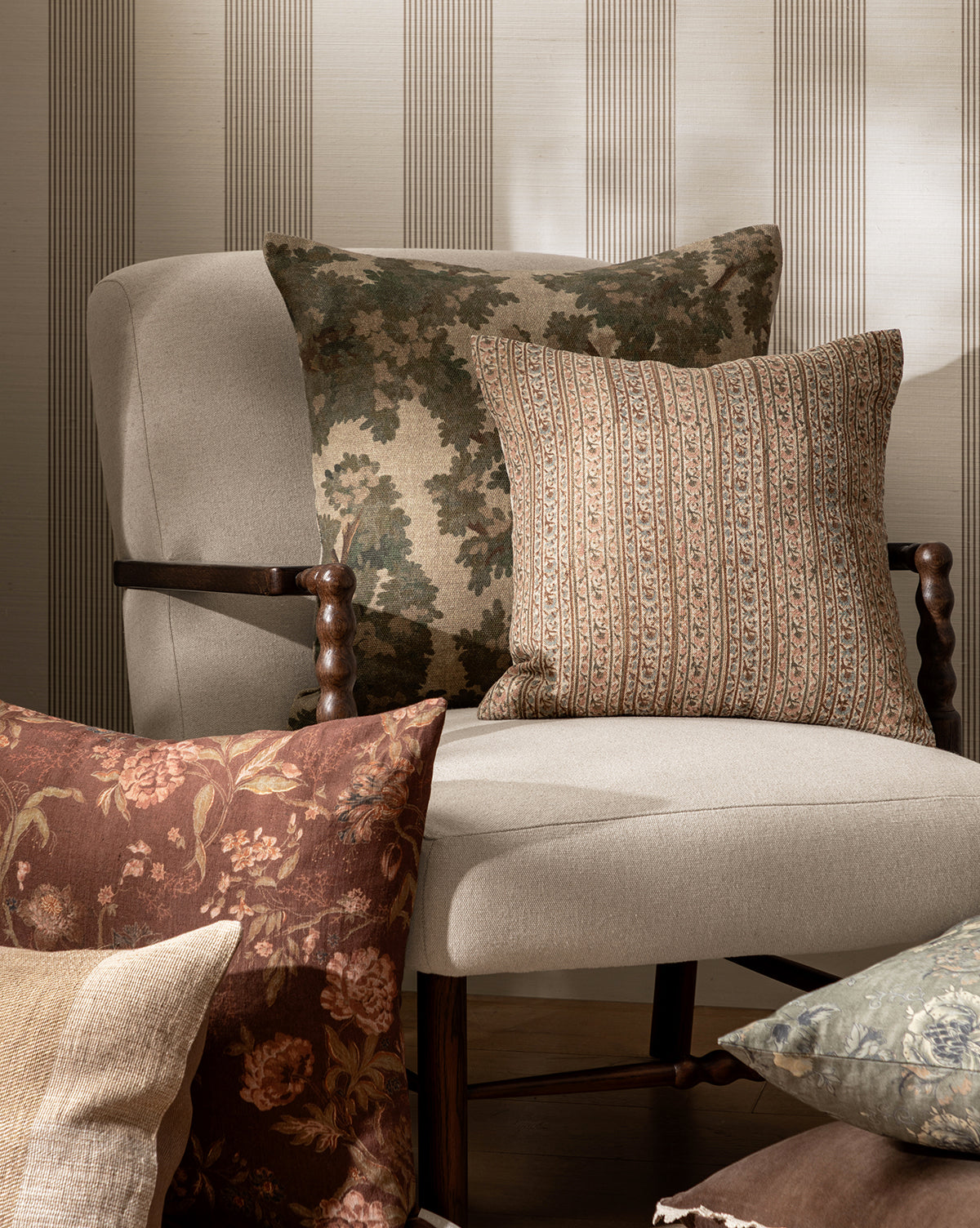 A beige upholstered chair holds two pillows—one adorned with the Wakefield Pillow Cover, featuring a leafy camouflage design, and another with stripes. In front, more floral and patterned pillows in neutral tones are artfully arranged by Vendixs