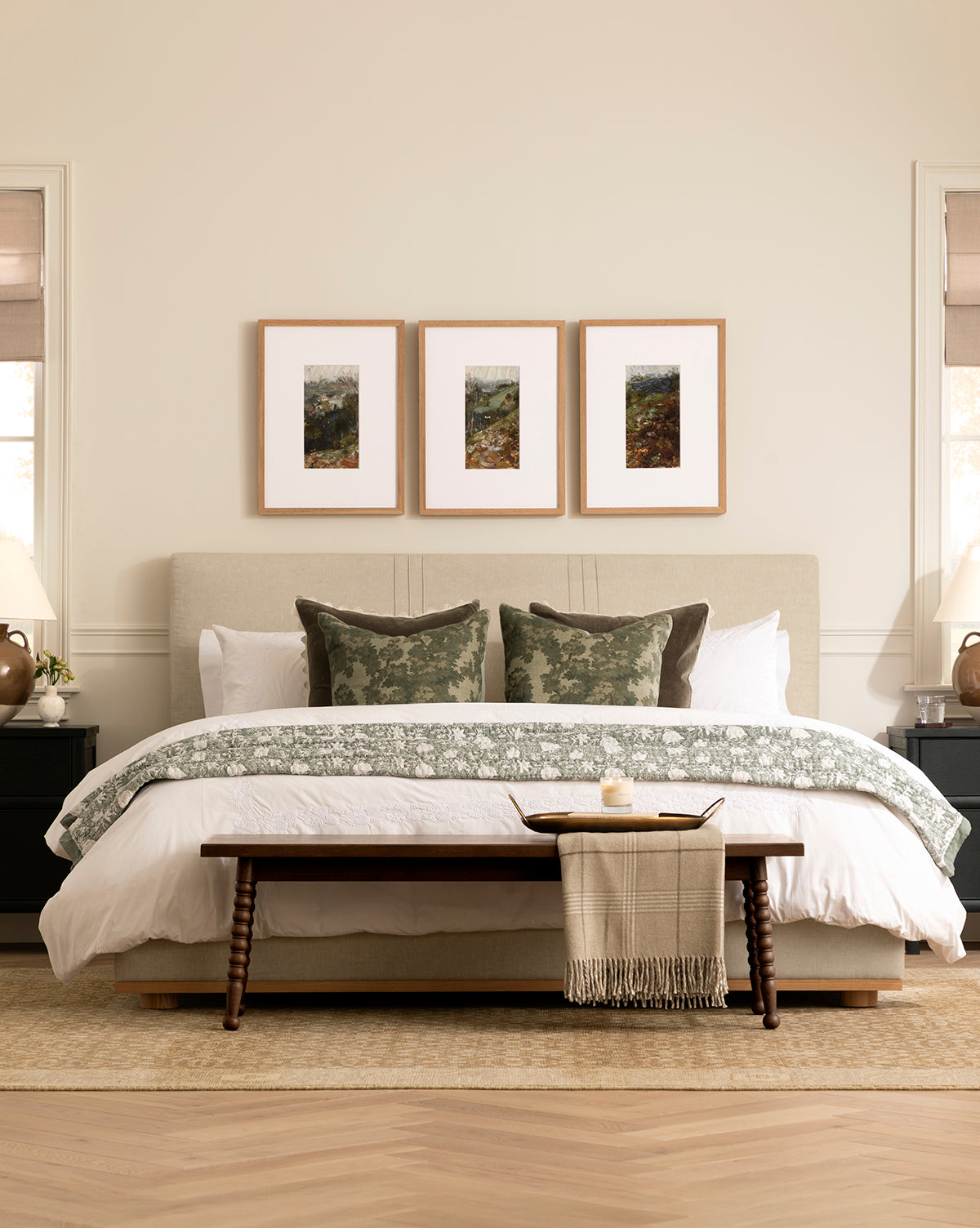 A neatly made bed with white and green bedding, three green pillows, and a Heathcliff Bench at the foot holding a tray and blanket; three framed artworks hang above, while bedside tables with lamps are on each side, by McGee & Co.
