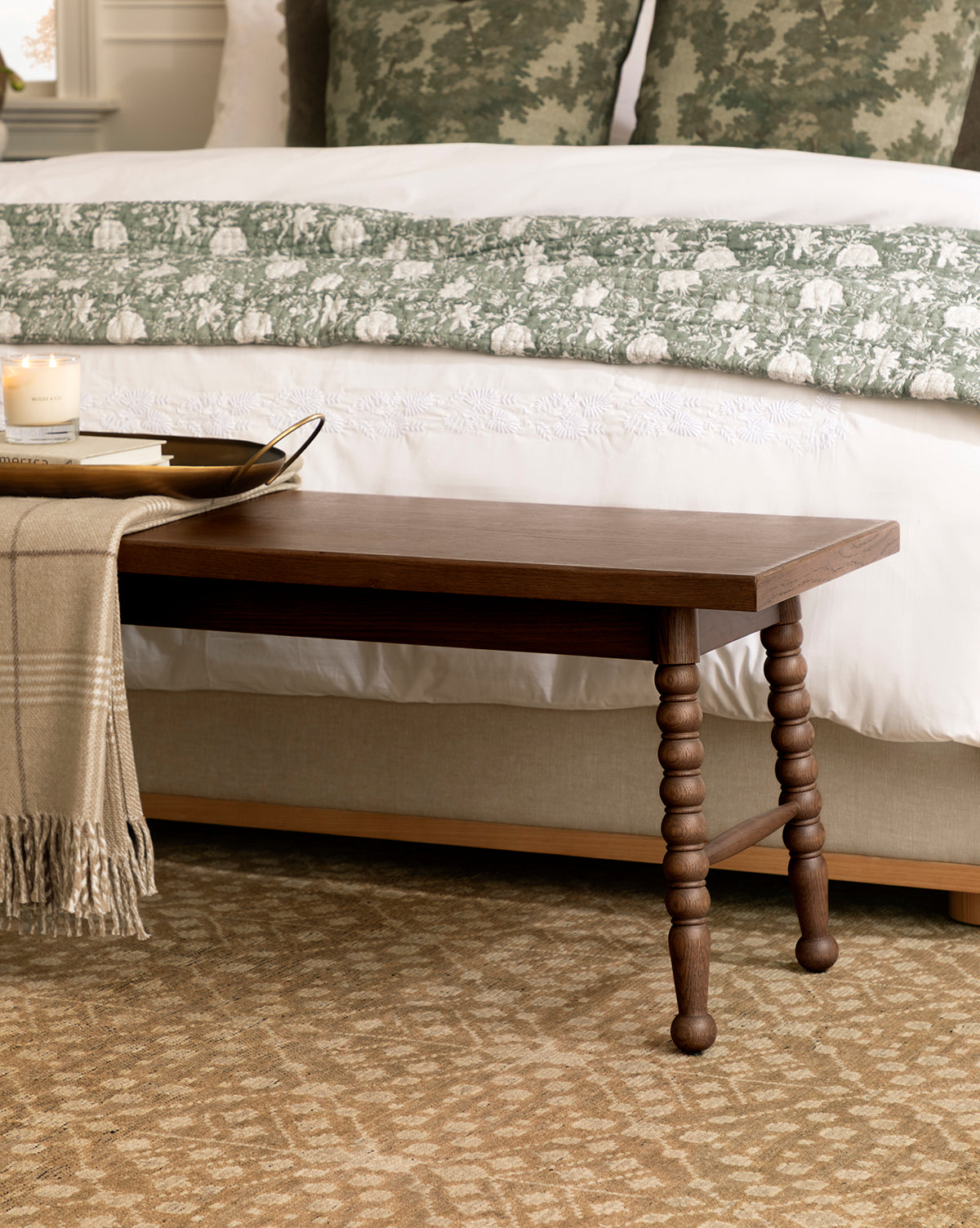 A wooden bench with turned legs sits at the foot of a bed with white and green patterned bedding, atop a Windsor Hand-Knotted Wool Rug; a beige throw blanket and a tray with a candle rest on the bench, by Vendixs