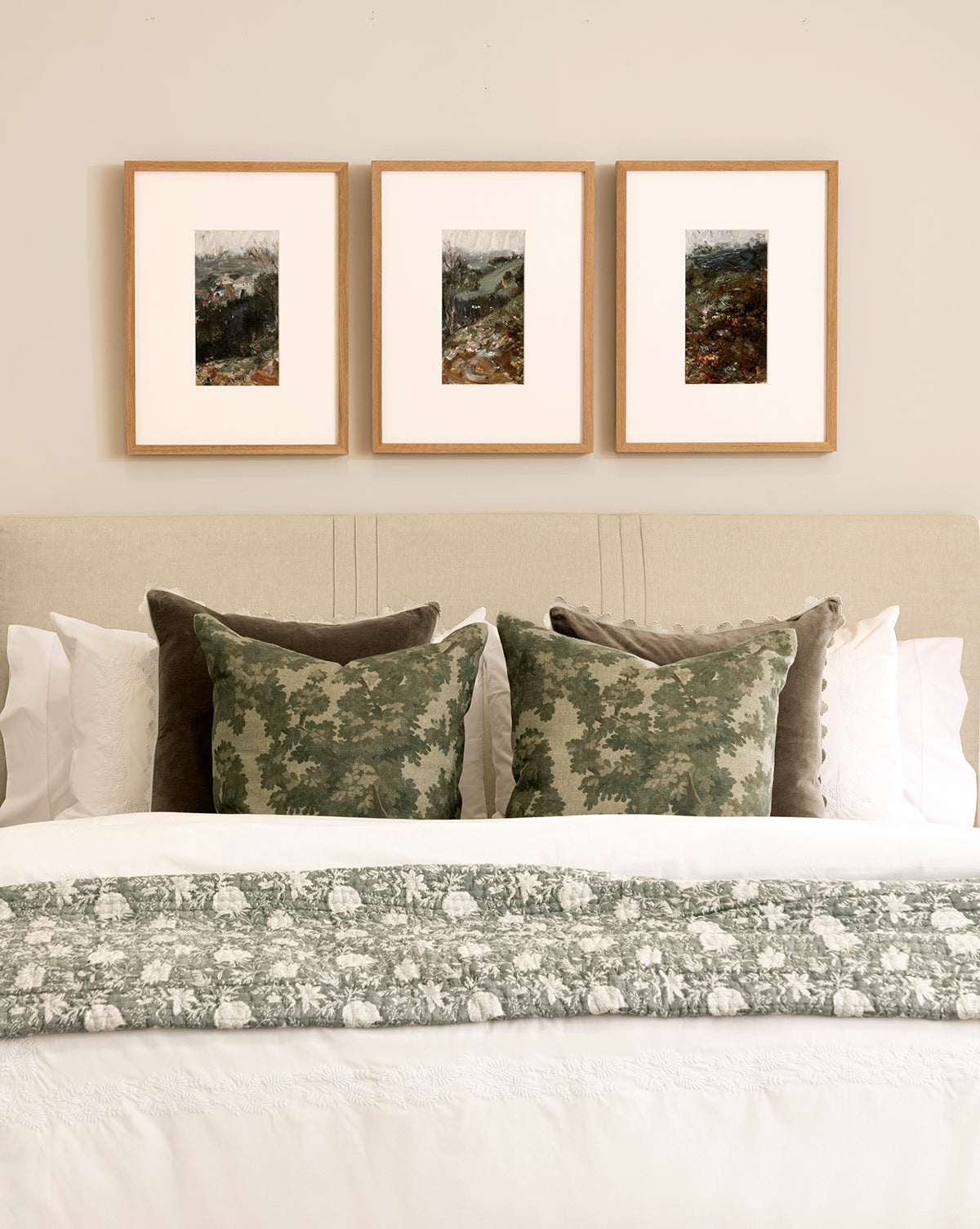 A neatly made bed with white linens and green pillows sits beneath A River Runs Through, a set of three framed abstract landscapes, adding striking wall art above the beige headboard on the light wall, by Vendixs