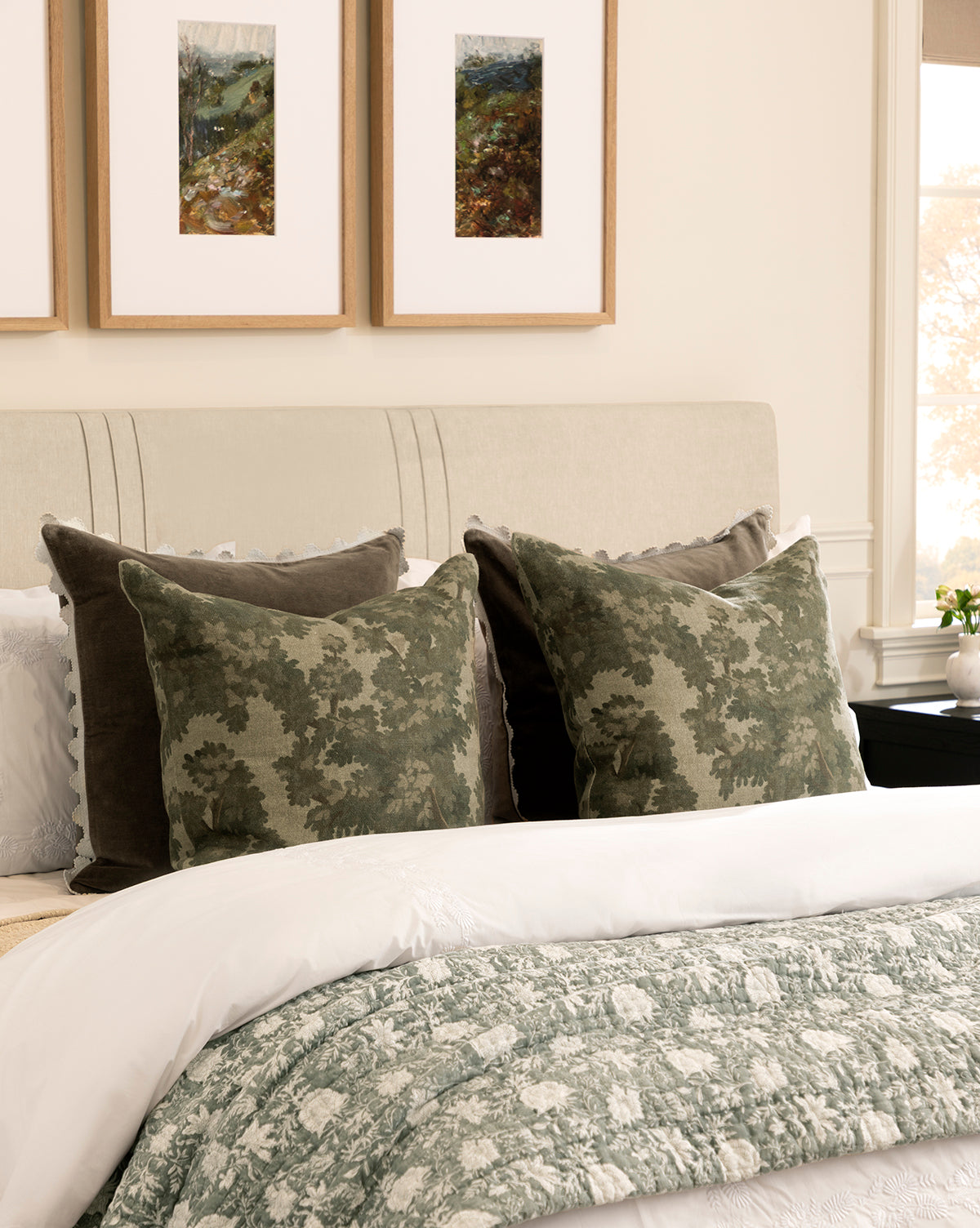 A neatly made bed features green and white floral bedding, two Lucetta Pillow Covers with a leafy pattern in earthy tones, two solid dark green pillows, and three framed landscape artworks above on a light-colored wall, by Vendixs