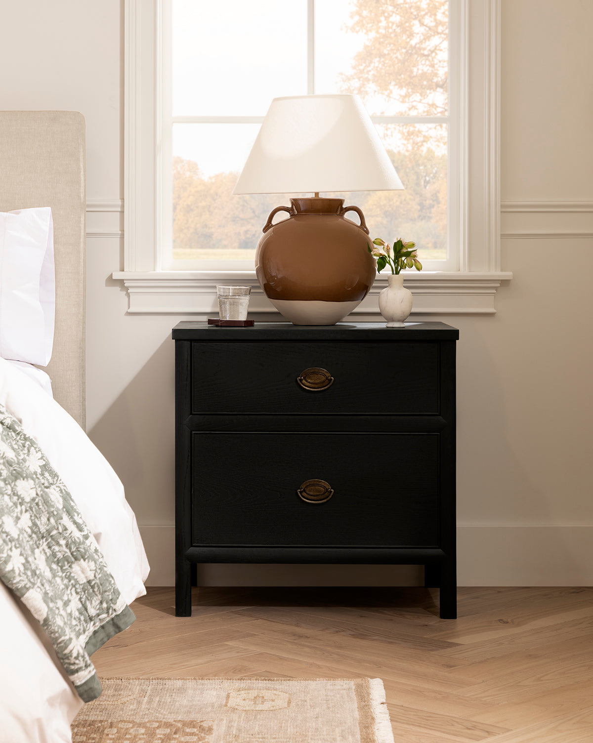 A black nightstand with two drawers stands by a bed. On top are a brown ceramic lamp with a white shade, a glass of water, the Fera Bud Vase holding greenery, and a book. Sunlight streams through the window in the background, by Vendixs