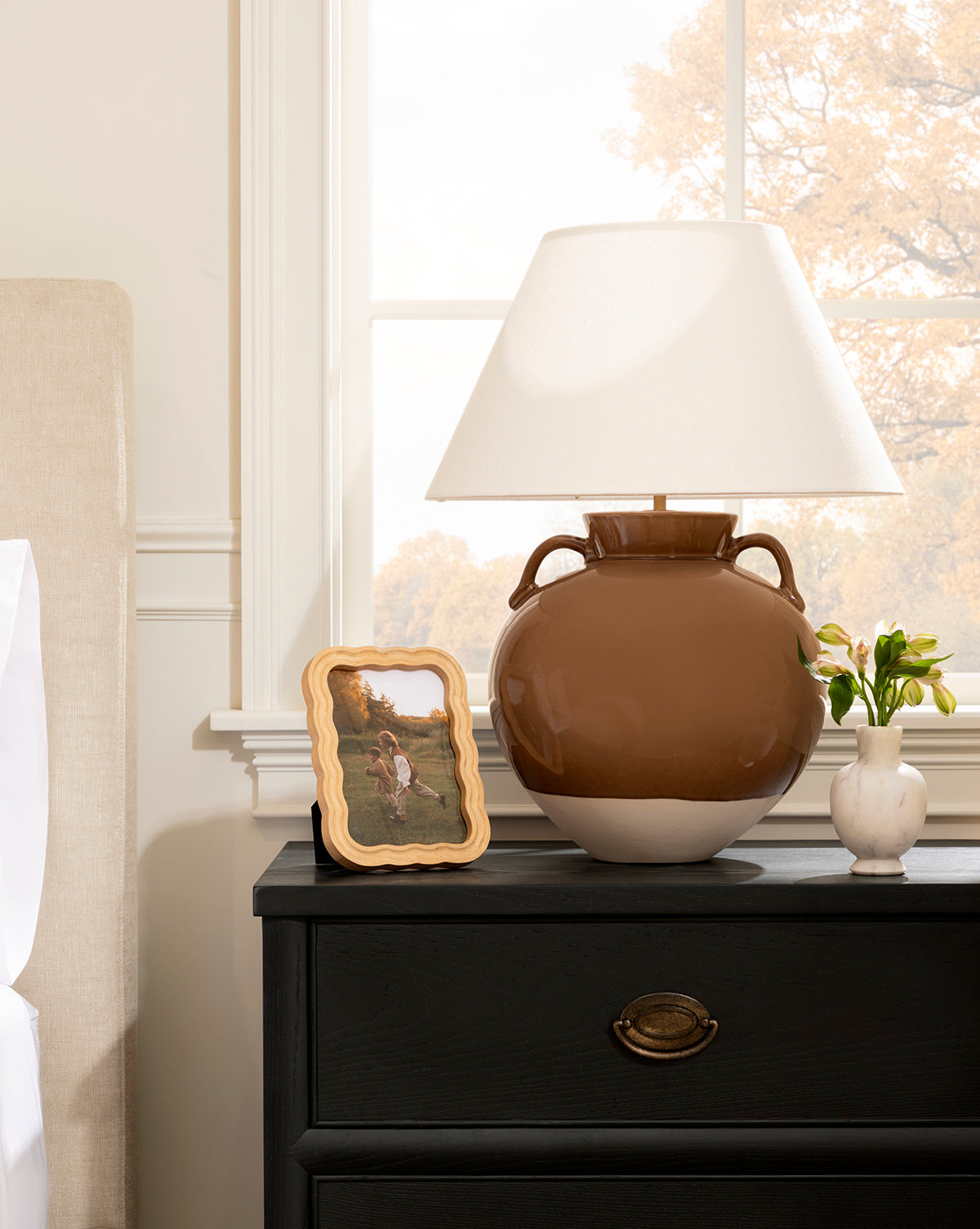 A natural rubberwood bedside table with a dark drawer displays a large brown lamp with a white shade, yellow flowers in a vase, and a photo in the Scalloped Wood Frame. The room includes a bed and window showing autumn trees outside, by Vendixs