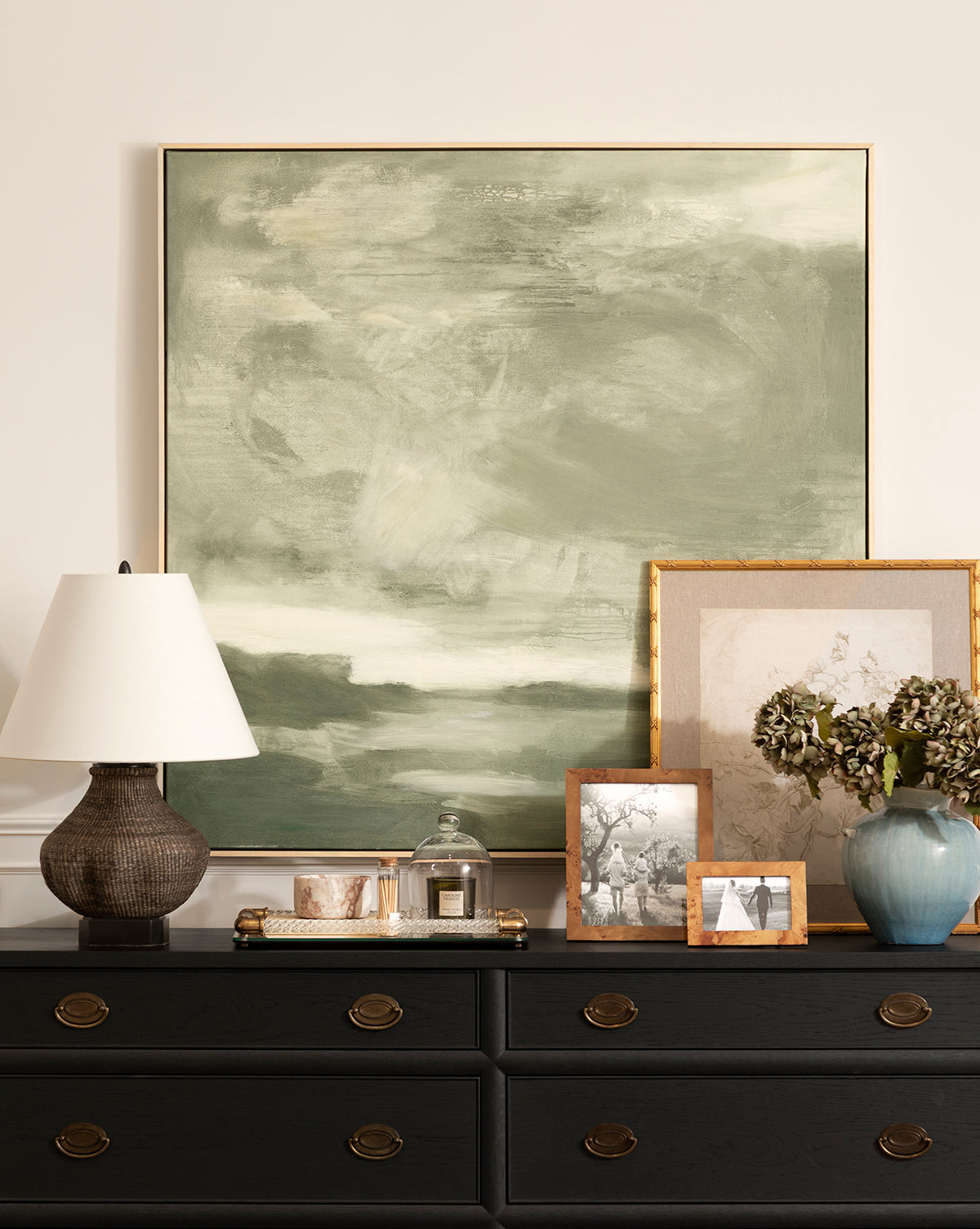 A dresser with round brass handles holds a textured lamp, a tray with a candle and framed photo, a blue vase featuring Feathery Floral, and grouped framed photos. Above hangs a large abstract green and white giclée on paper, by Vendixs