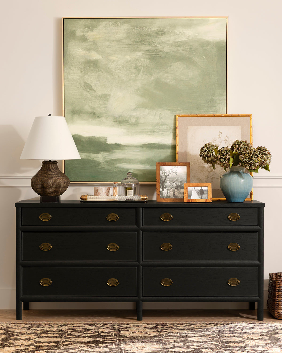 The Edward Dresser in Black Ash with brass knobs provides chic bedroom storage, adorned with a textured lamp, tray, glass jar, framed photos, vase of dried flowers, and two large overlapping art pieces above a patterned rug, by Vendixs