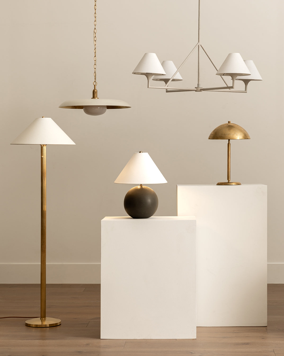 The Townes Floor Lamp in gold reeded brass, a black table lamp, and a gold desk lamp stand on white pedestals; above are a white linen-shade chandelier and a gold pendant, all set against a neutral backdrop, by Vendixs