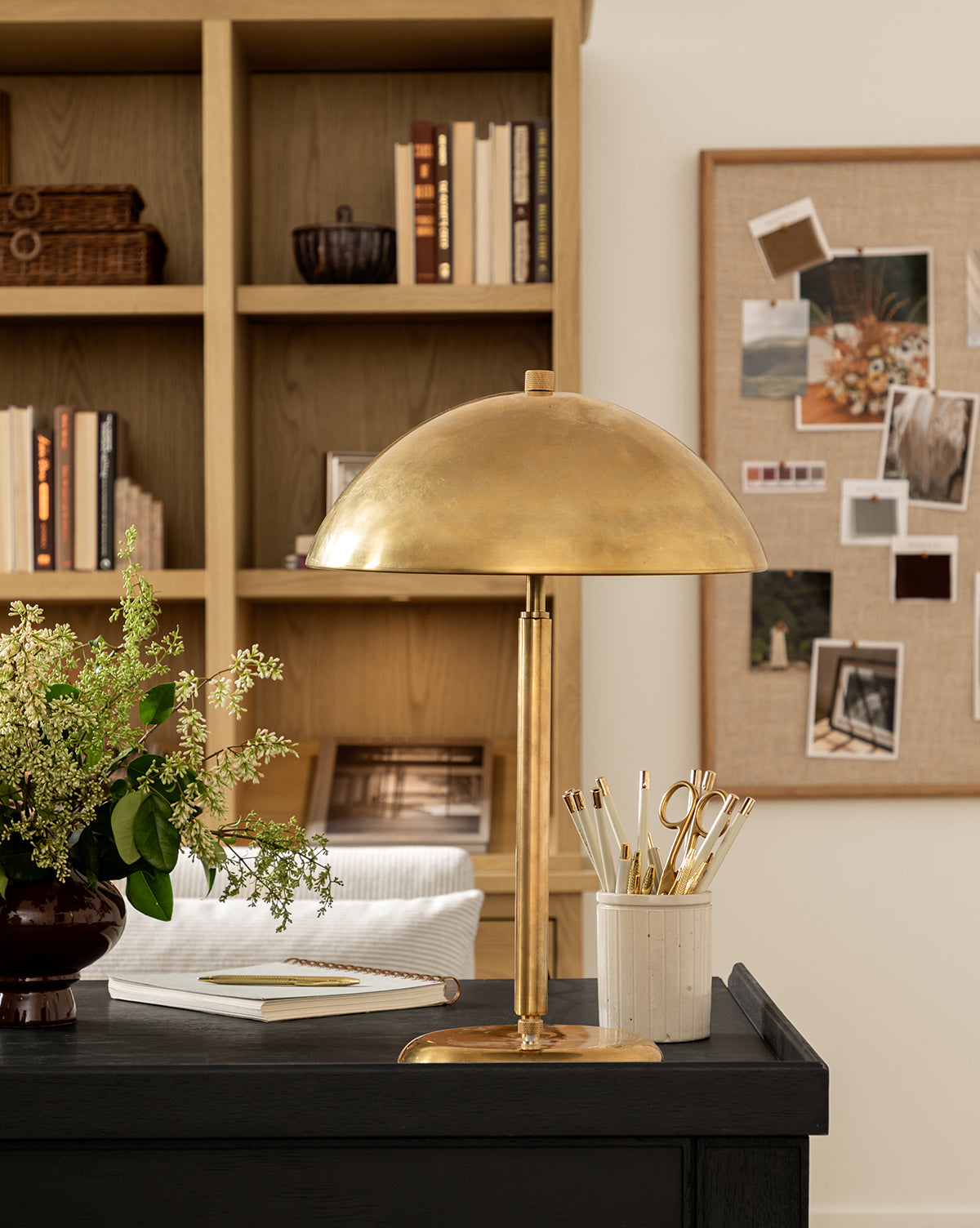 The Aldorno Desk Table Lamp sits on a black desk with gold pens, a plant, and a notebook. Behind it, wooden shelves hold books and baskets, while a bulletin board displays photos—perfect for stylish living room lighting, by Vendixs