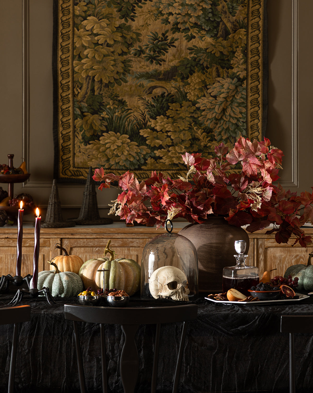 A dark, elegant table is decorated for autumn with pumpkins, a skull under a glass dome, candles, treats, and a Faux Autumn Grapevine Stem in a vase. An ornate tapestry with foliage hangs on the wall behind this seasonal arrangement, by Vendixs